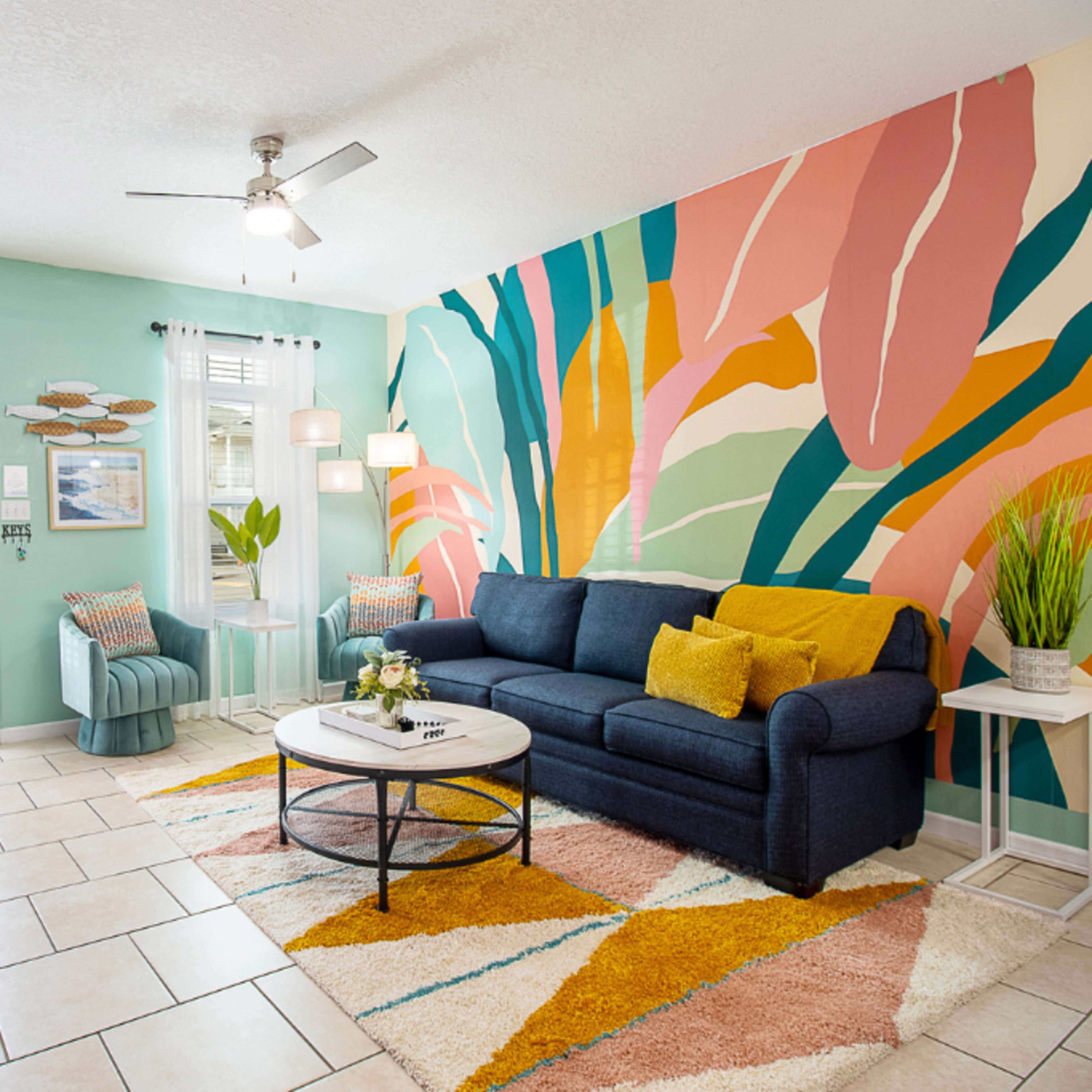 A brightly decorated living room featuring a blue sofa, colorful patterned rug, and a large tropical mural on the wall.