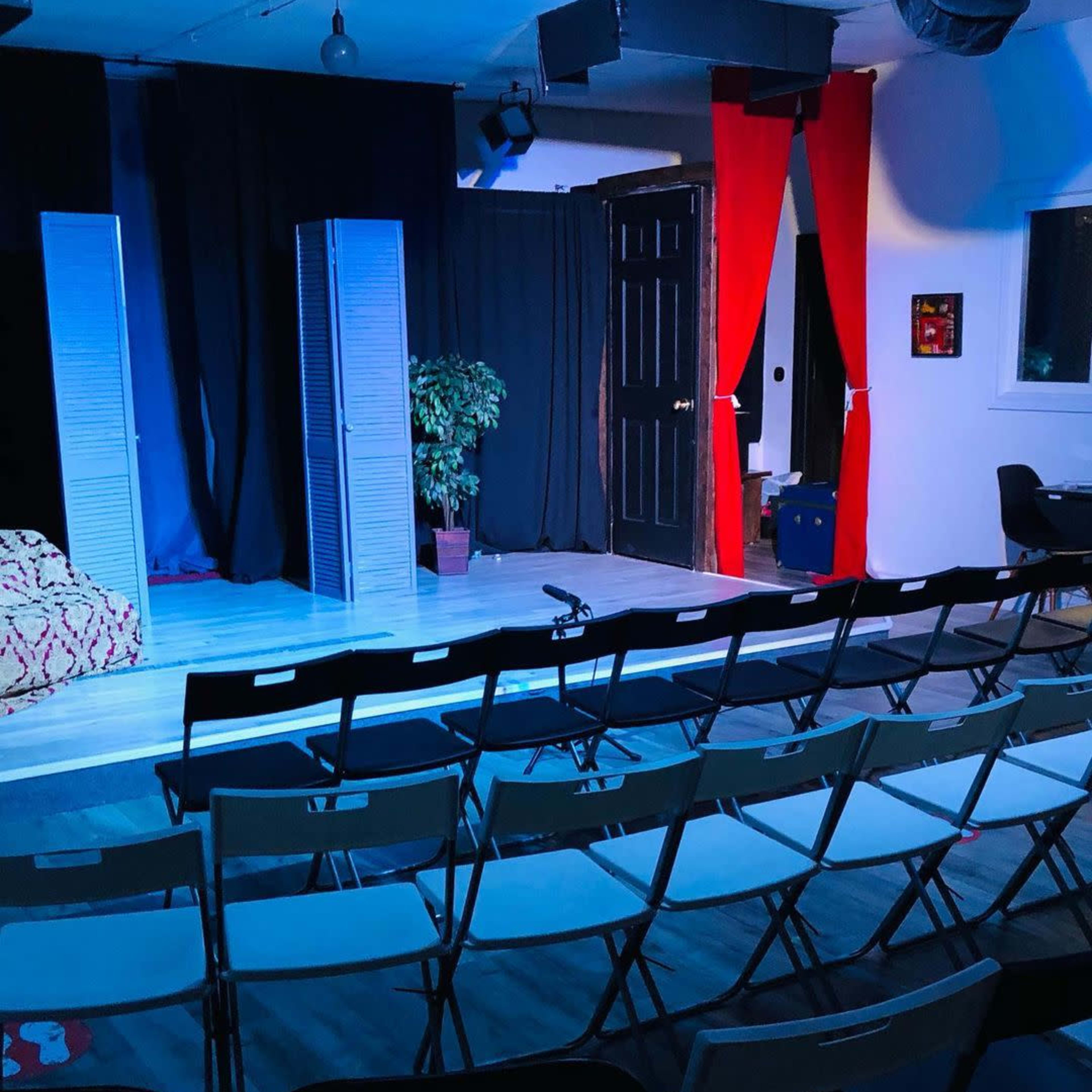 A theater stage with two folding screens, a plant in a pot, red curtains, and rows of empty black chairs arranged for an audience.