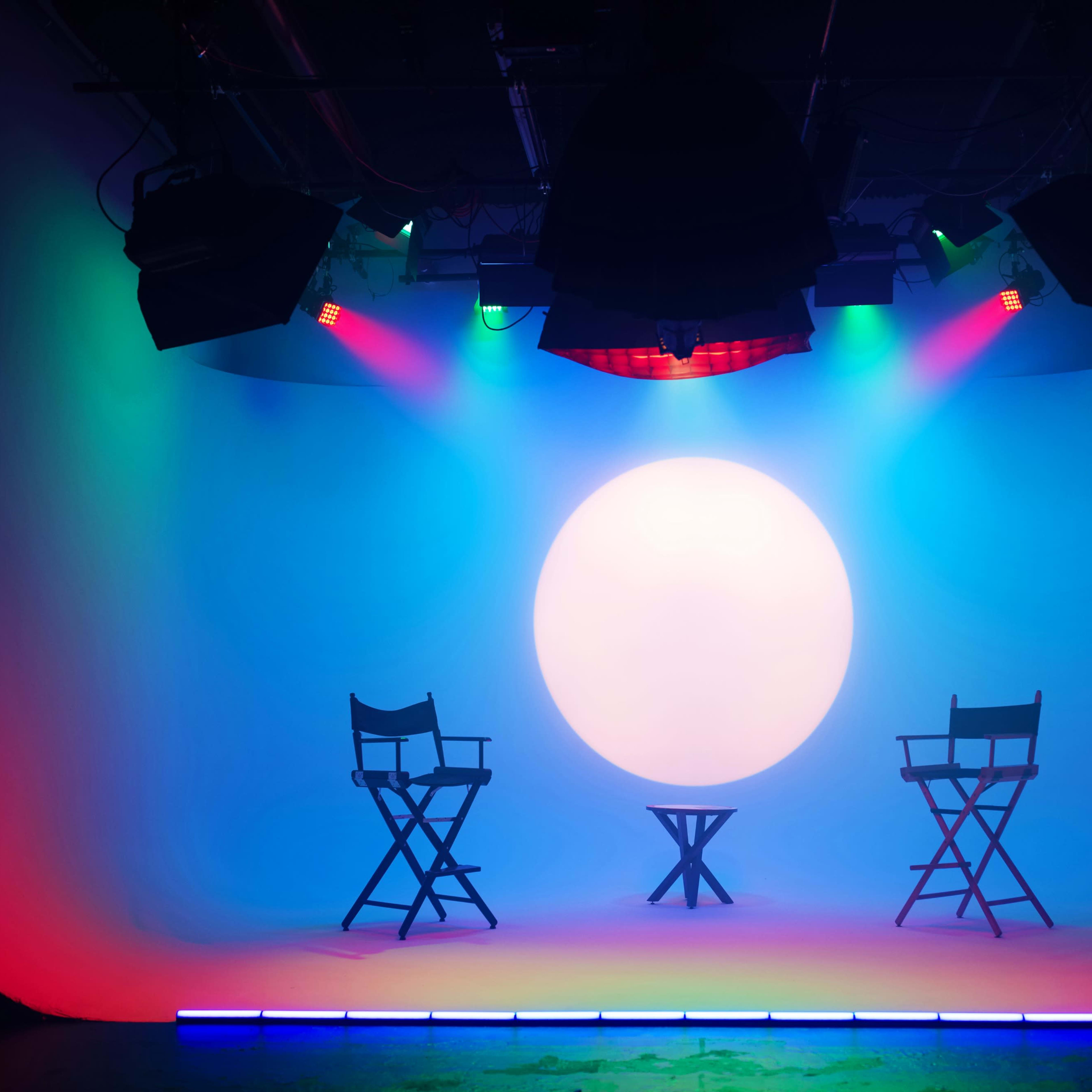 A minimalist studio setup with two director's chairs and a small table, illuminated by colorful stage lights against a large circular backdrop.
