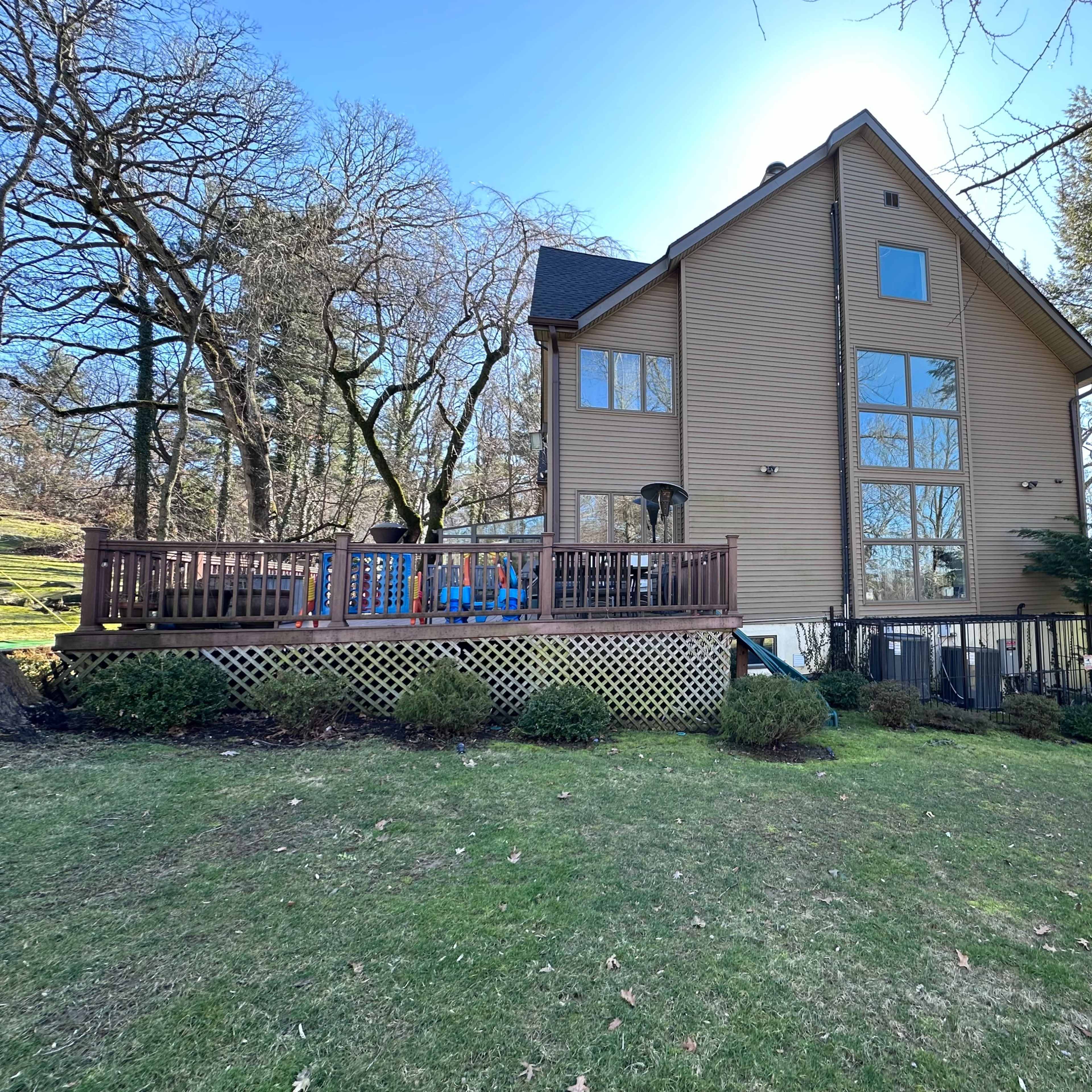 A large two-story house with a wooden deck and multiple windows is situated on a grassy lawn surrounded by trees.