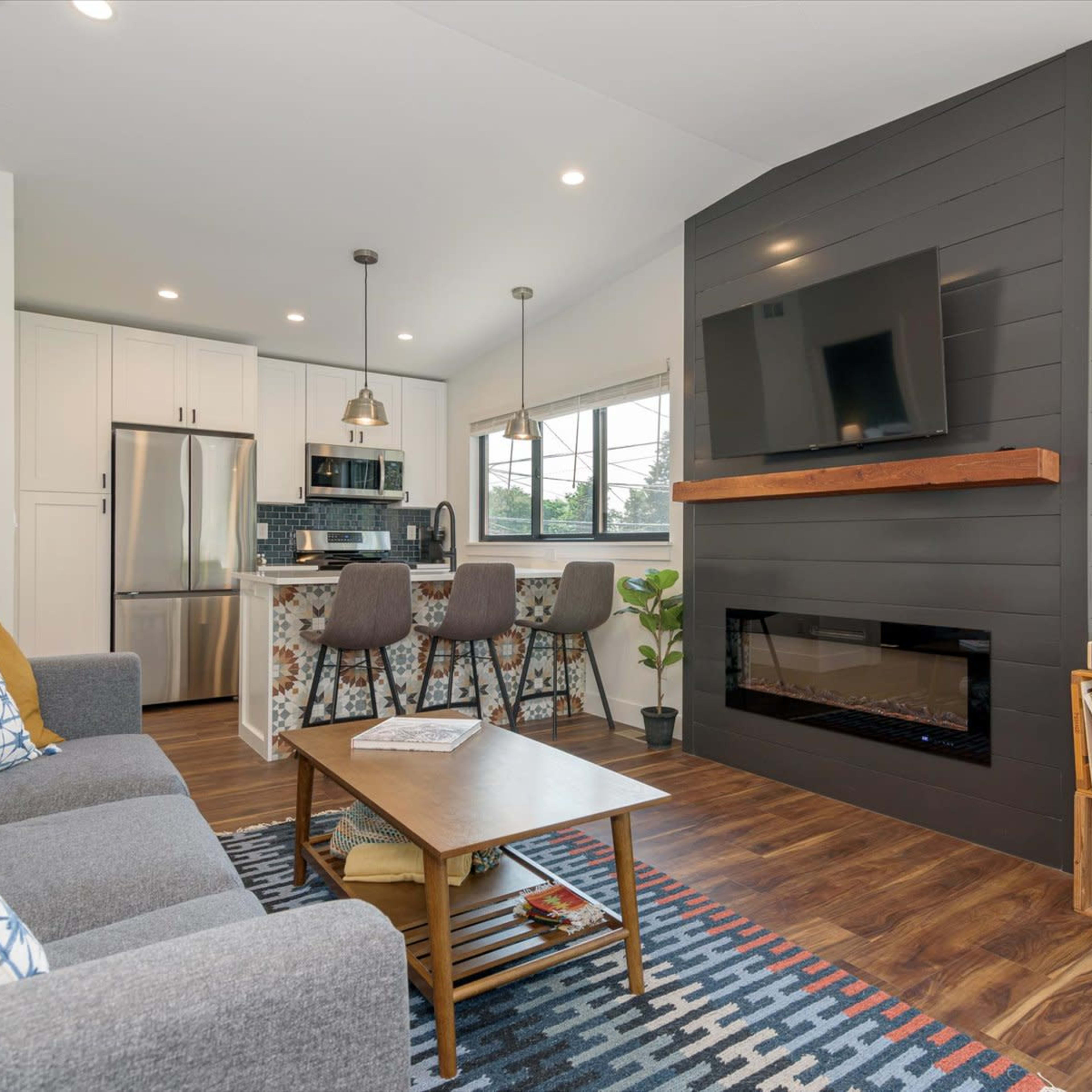 A modern living room and kitchen area with a seating arrangement, a large television mounted on a wall, and a fireplace below it.