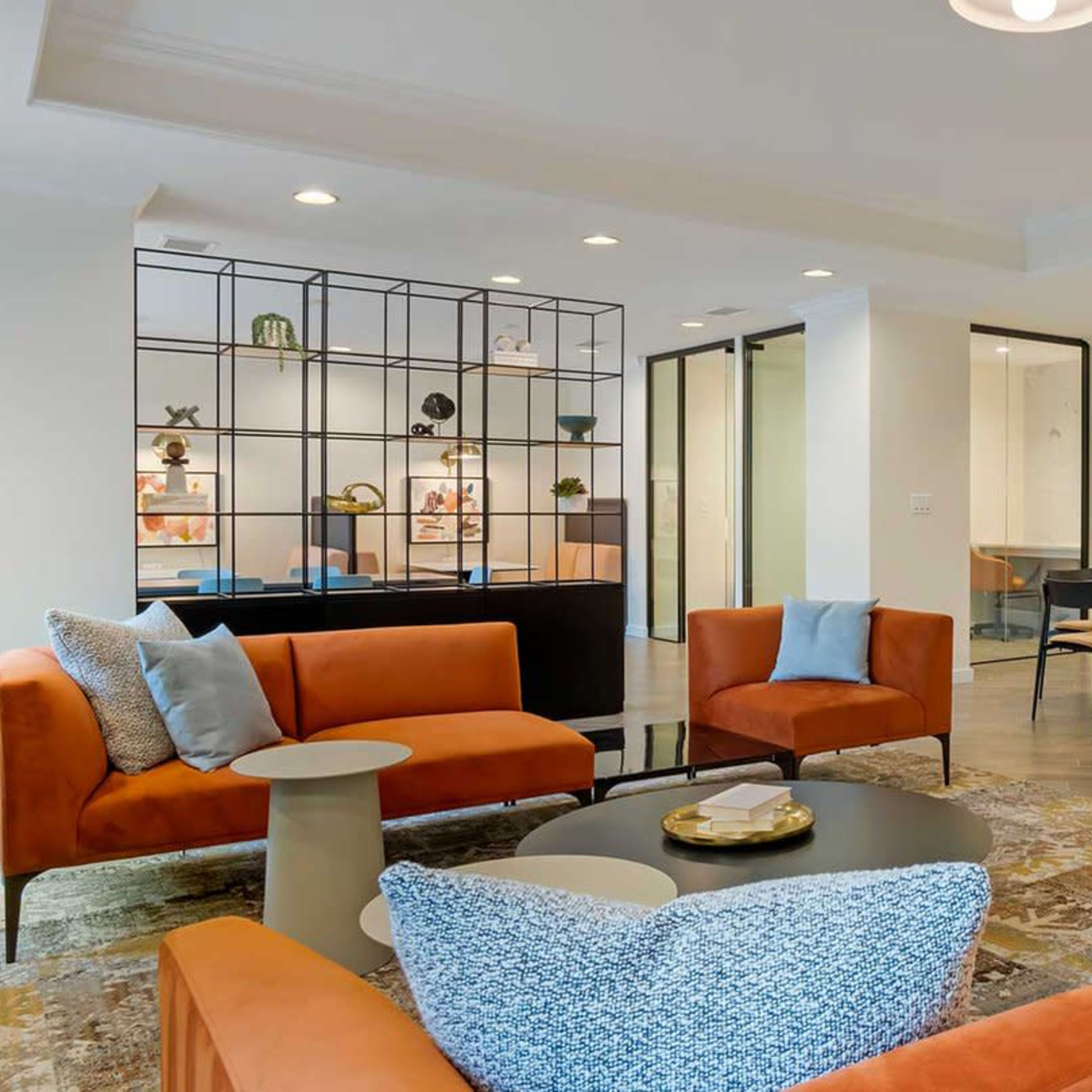 A modern lounge area featuring orange sofas, a coffee table, and a decorative shelving unit, with a dining area visible in the background.