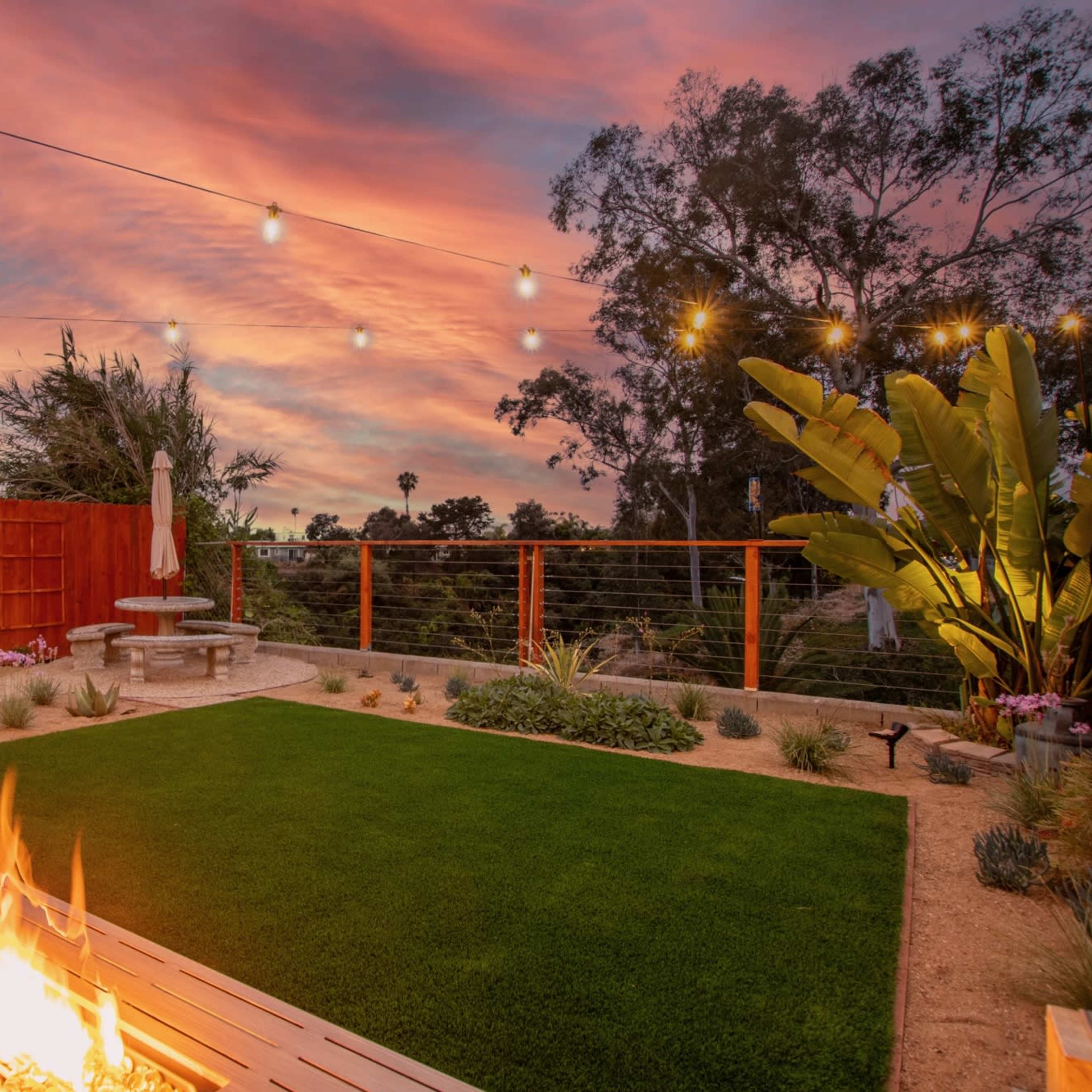 A landscaped backyard with a fire pit, green grass, and decorative string lights above at sunset.