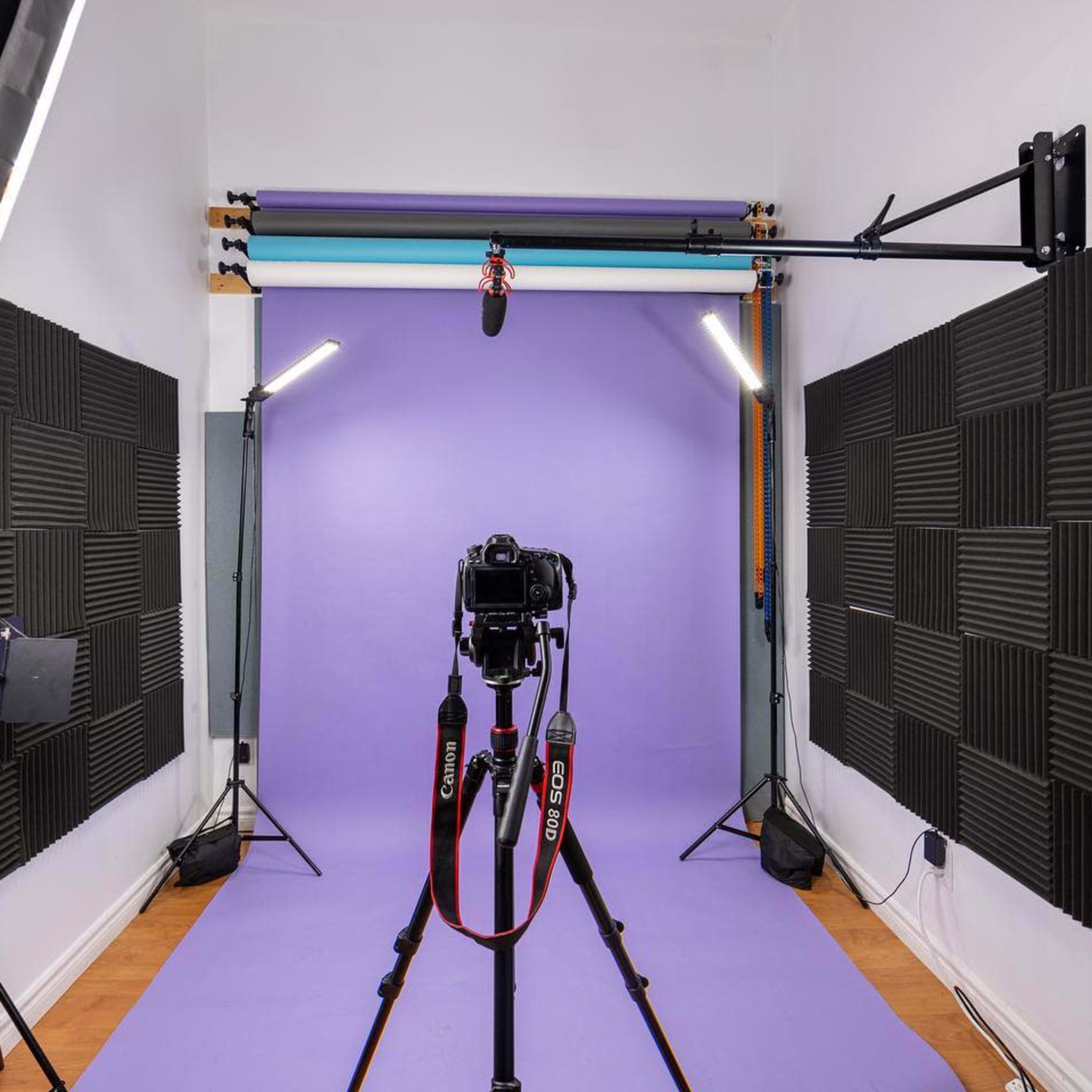 A photography studio features a camera on a tripod facing a purple backdrop, surrounded by various lighting equipment.