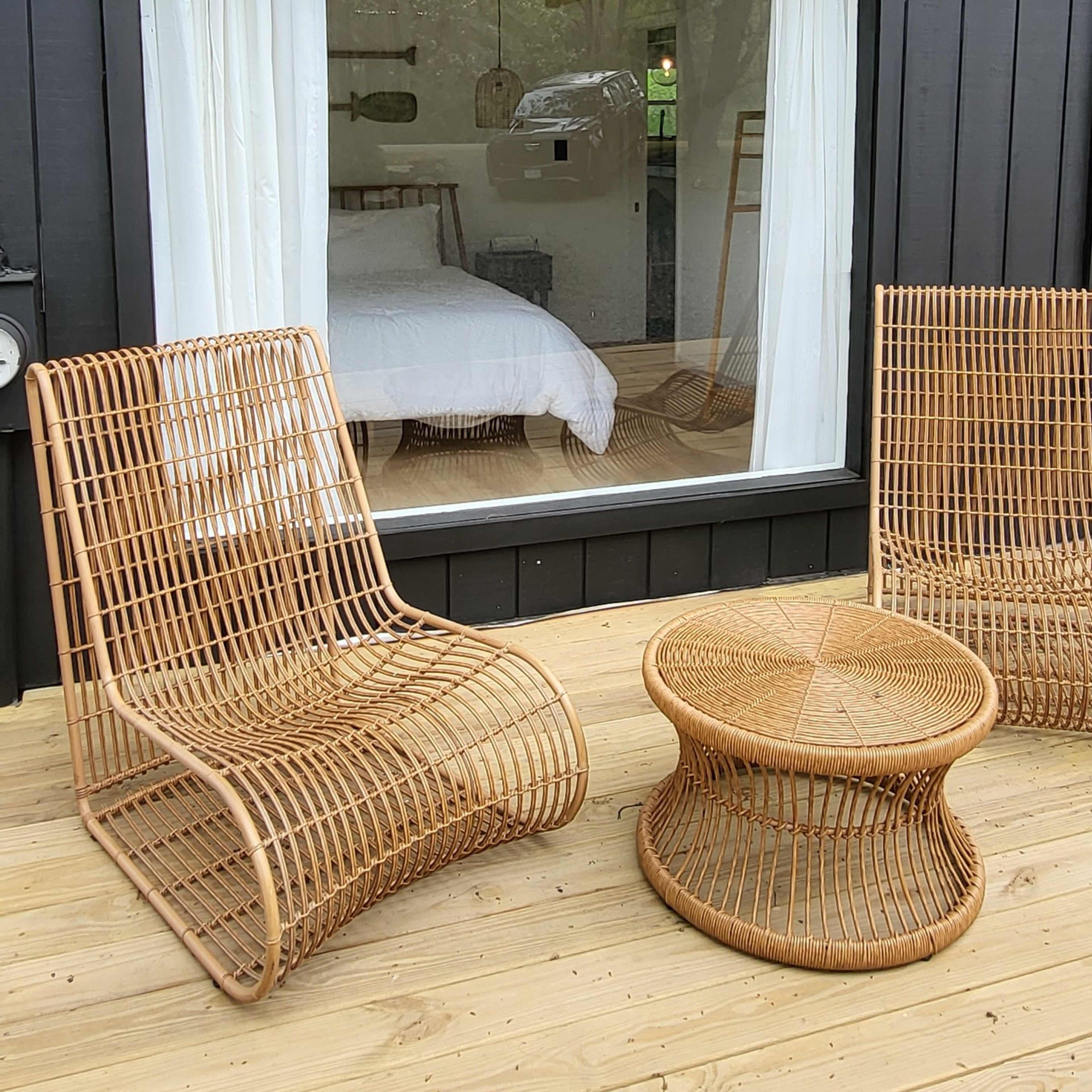 A deck area with two woven chairs and a round table, overlooking a window that reveals a bedroom with a bed and natural light.