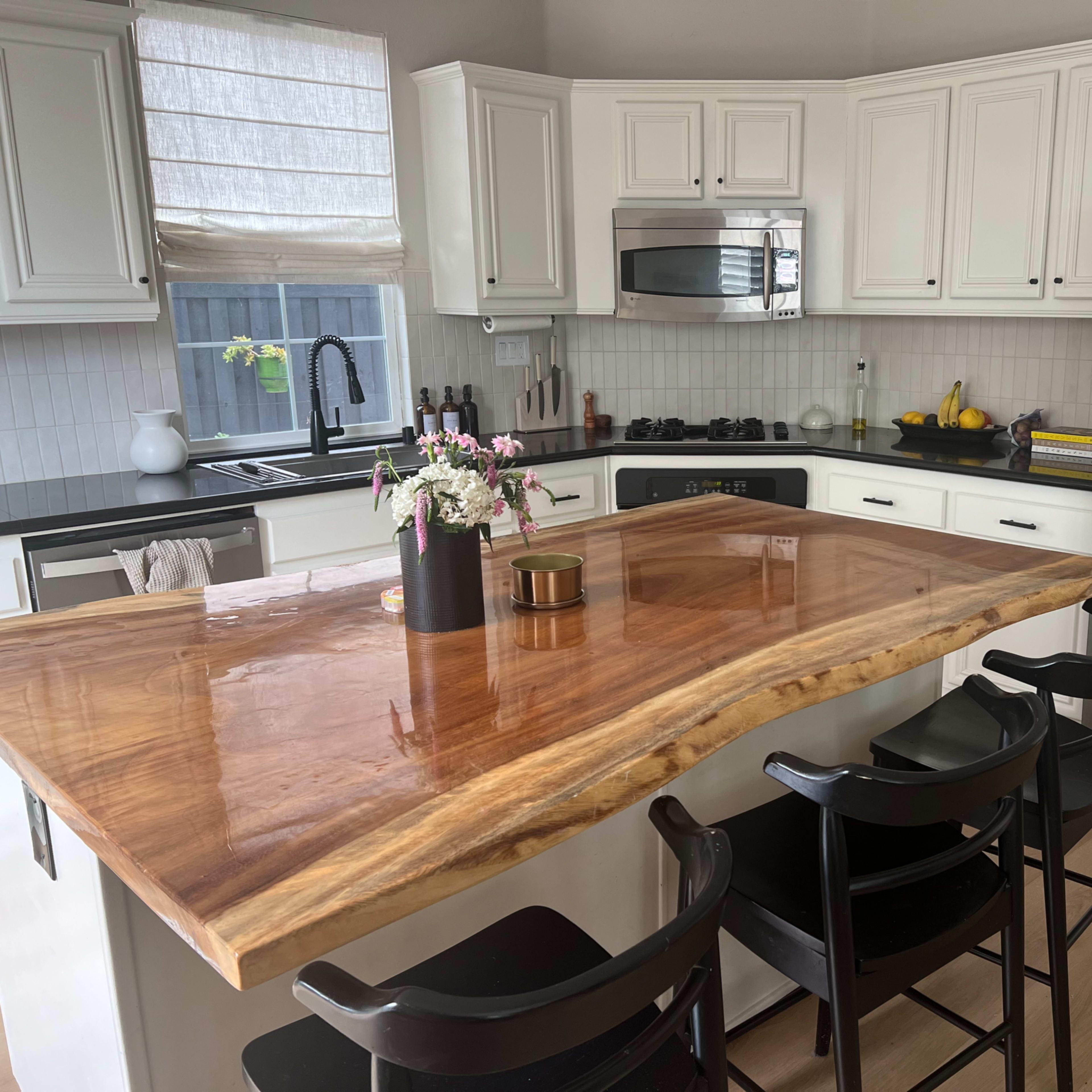 A modern kitchen features white cabinetry, a large wooden island counter, black chairs, and stainless steel appliances.
