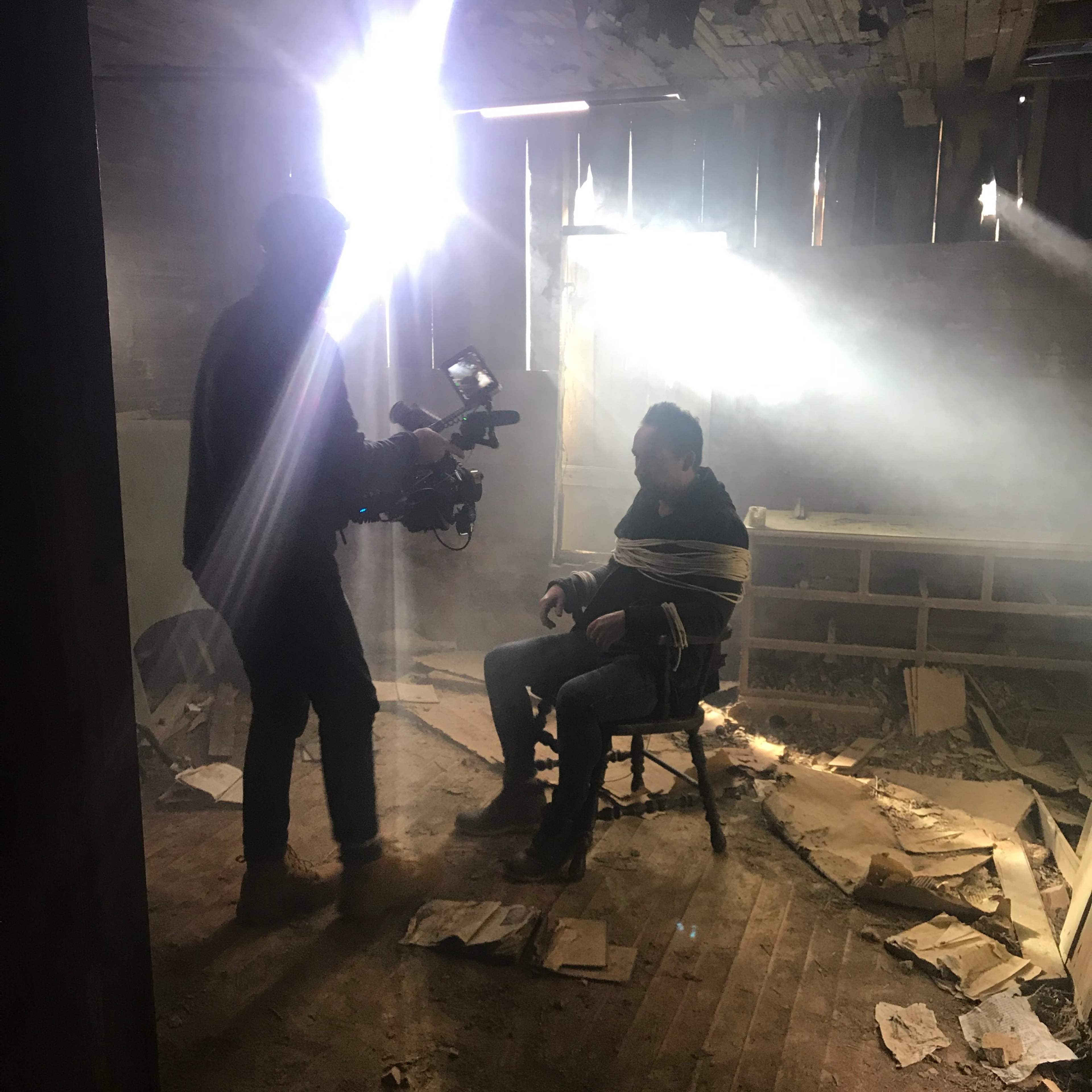 A man is tied to a chair in a dimly lit room while a person uses a camera to film him.