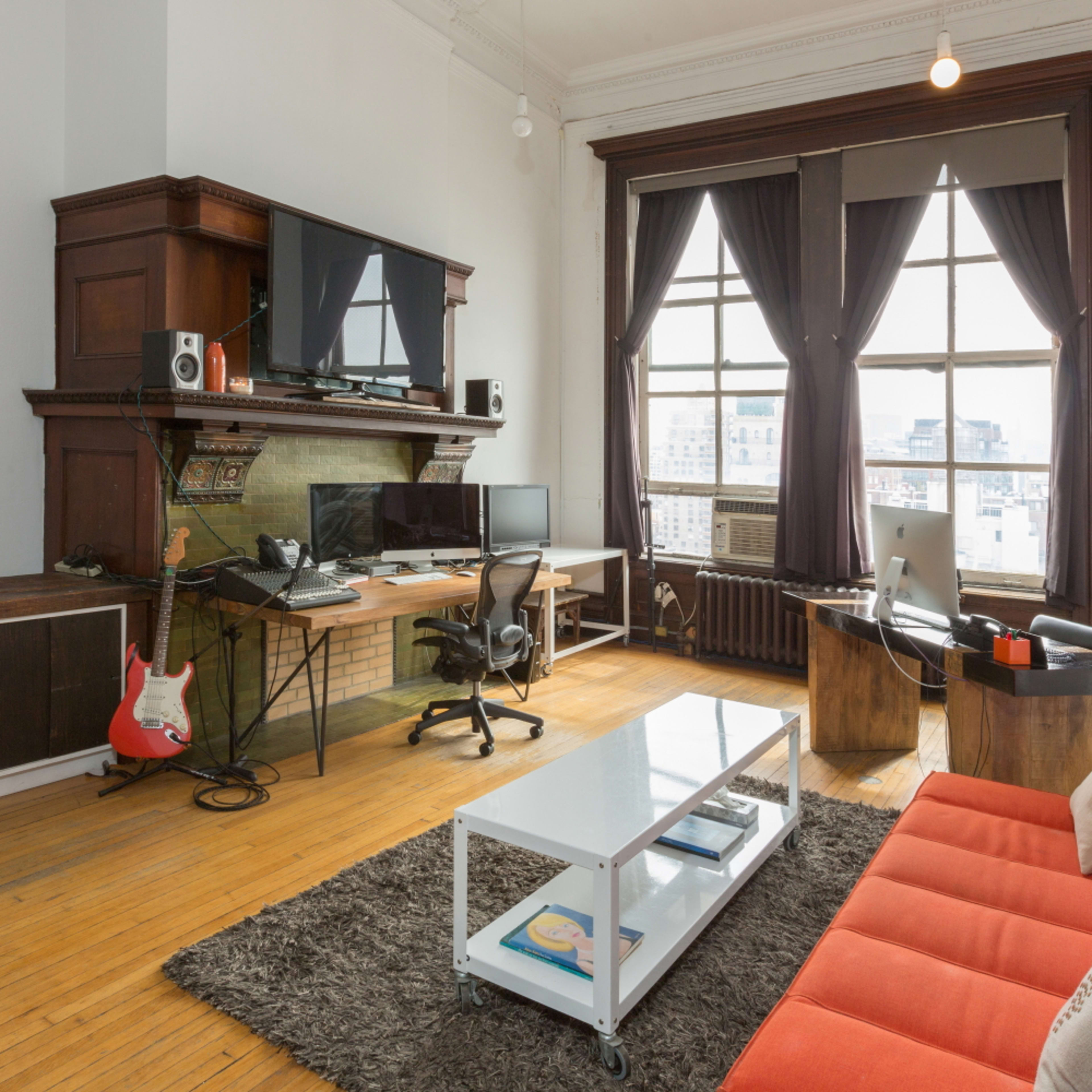 A bright, modern living space features a red sofa, a coffee table, and a workspace with two computers, all set against a backdrop of large windows and elegant curtains.