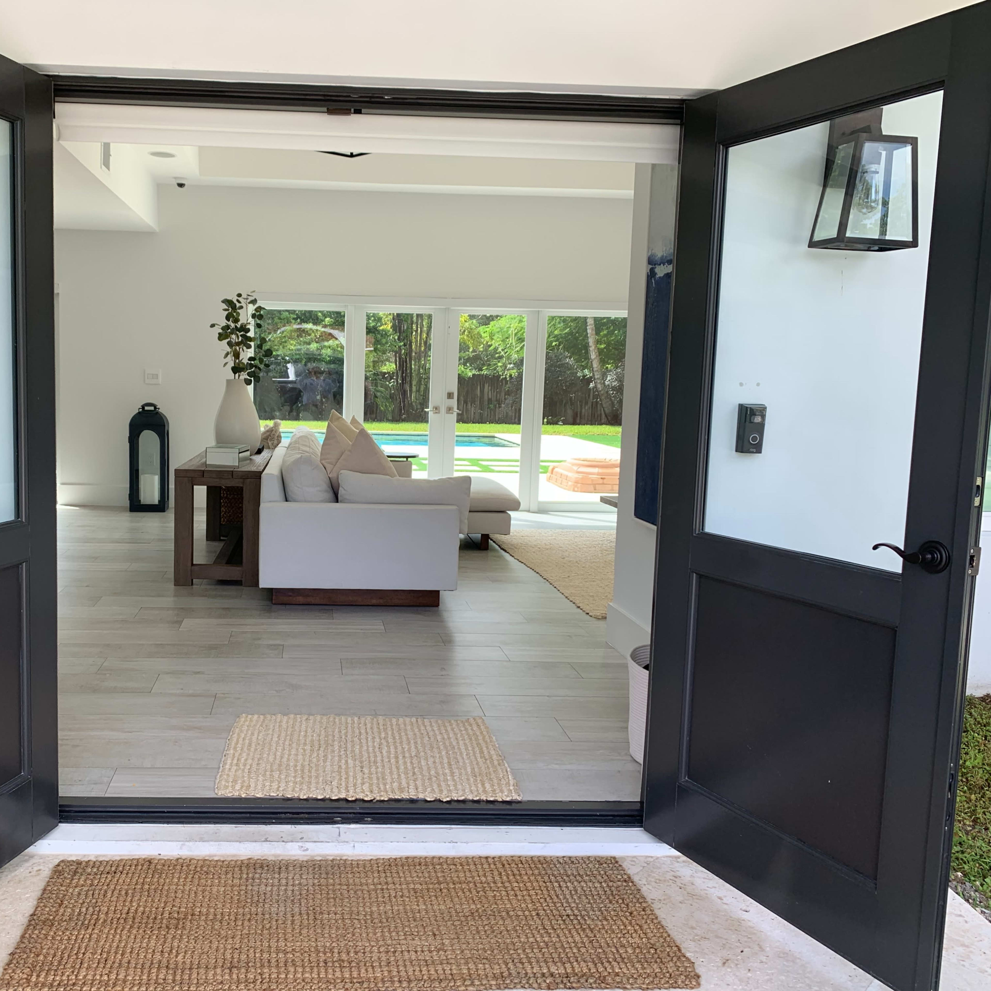 An open double door leading into a bright living space with a sofa and large windows that overlook an outdoor area.