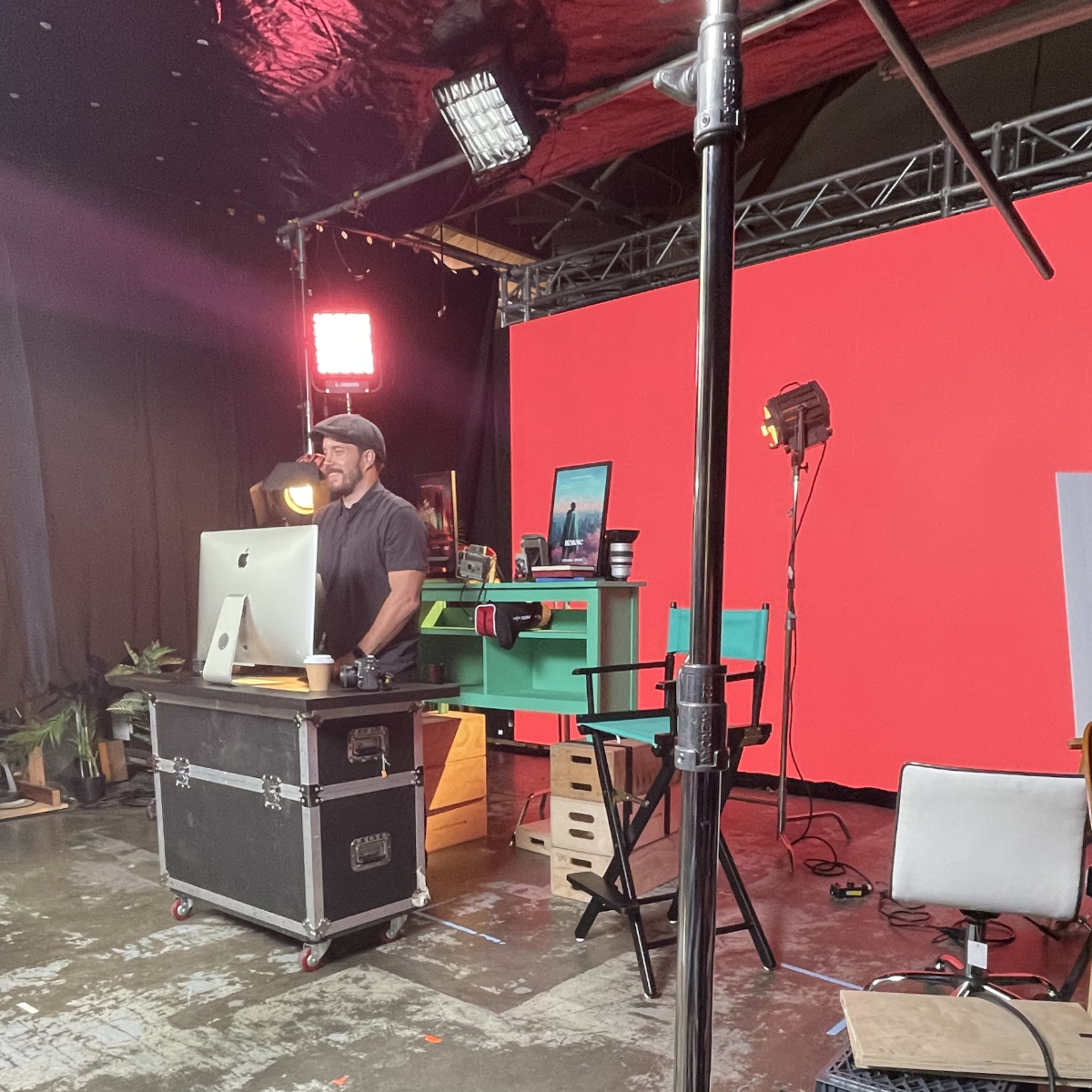 A person stands at a desk with a computer in a studio filled with lighting equipment and a bright red backdrop.
