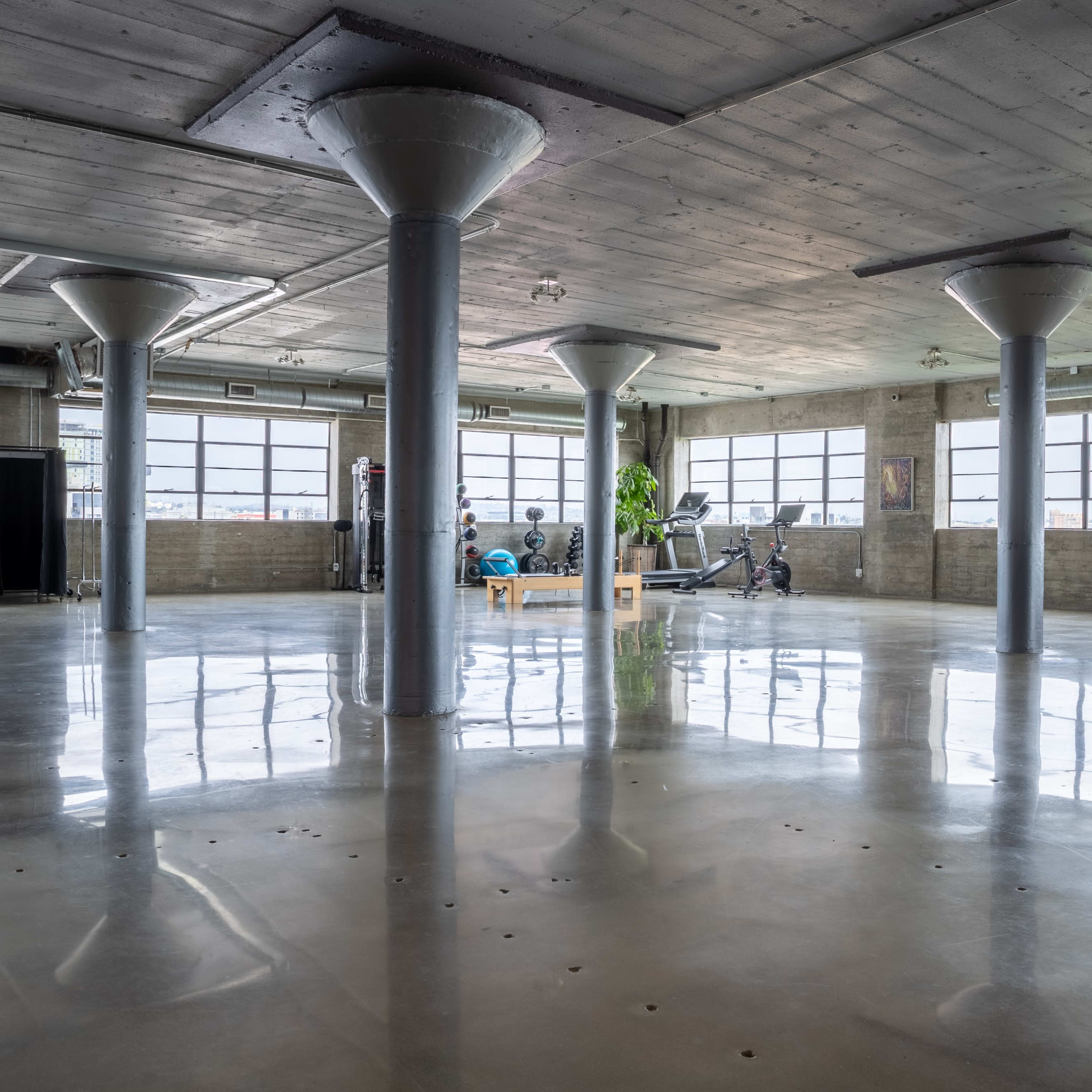 An open, industrial-style room features polished concrete floors, large windows, and fitness equipment with people exercising.