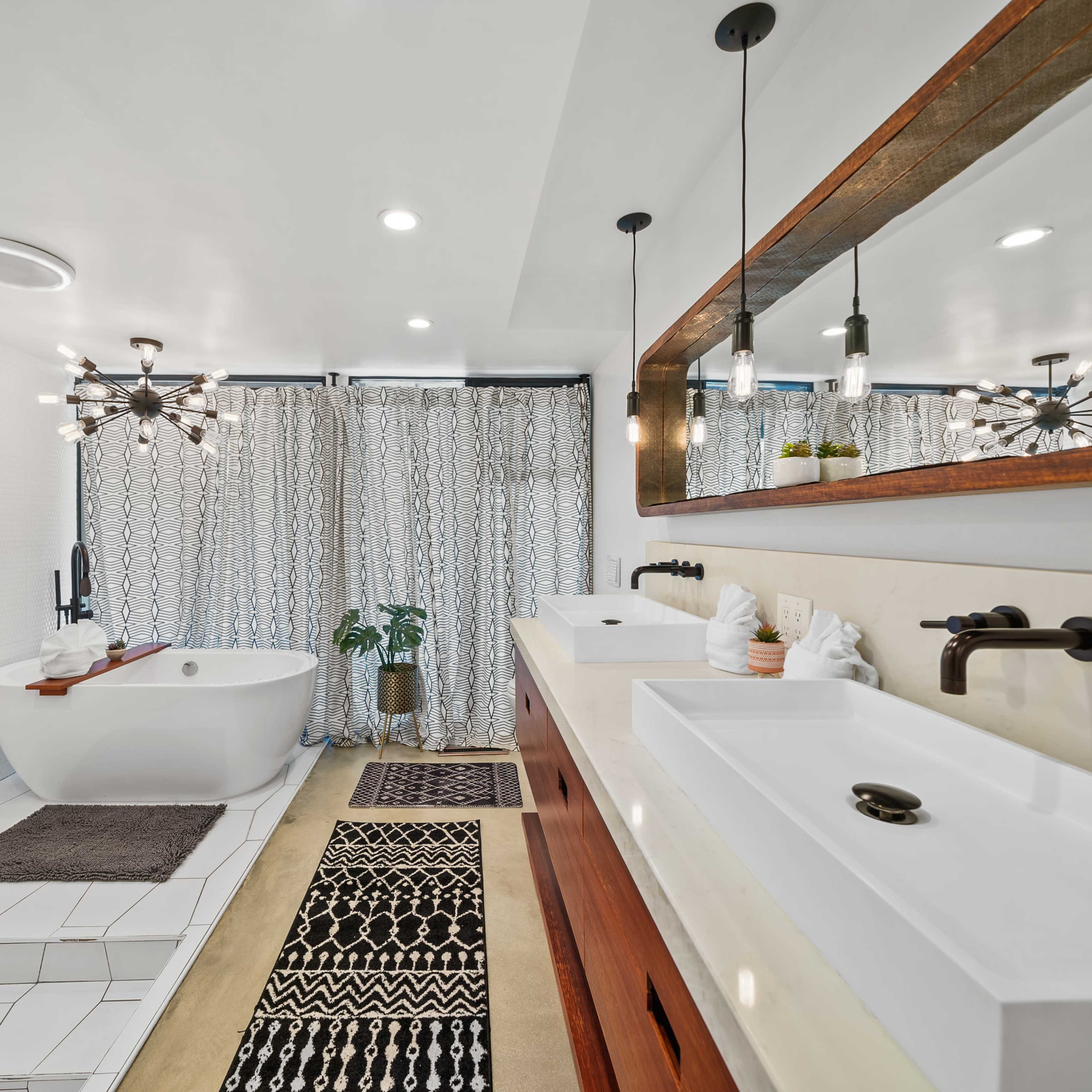 The bathroom features a freestanding tub, dual sinks with modern faucets, and a decorative rug under a large mirror, with light fixtures and a window curtain adding texture to the space.