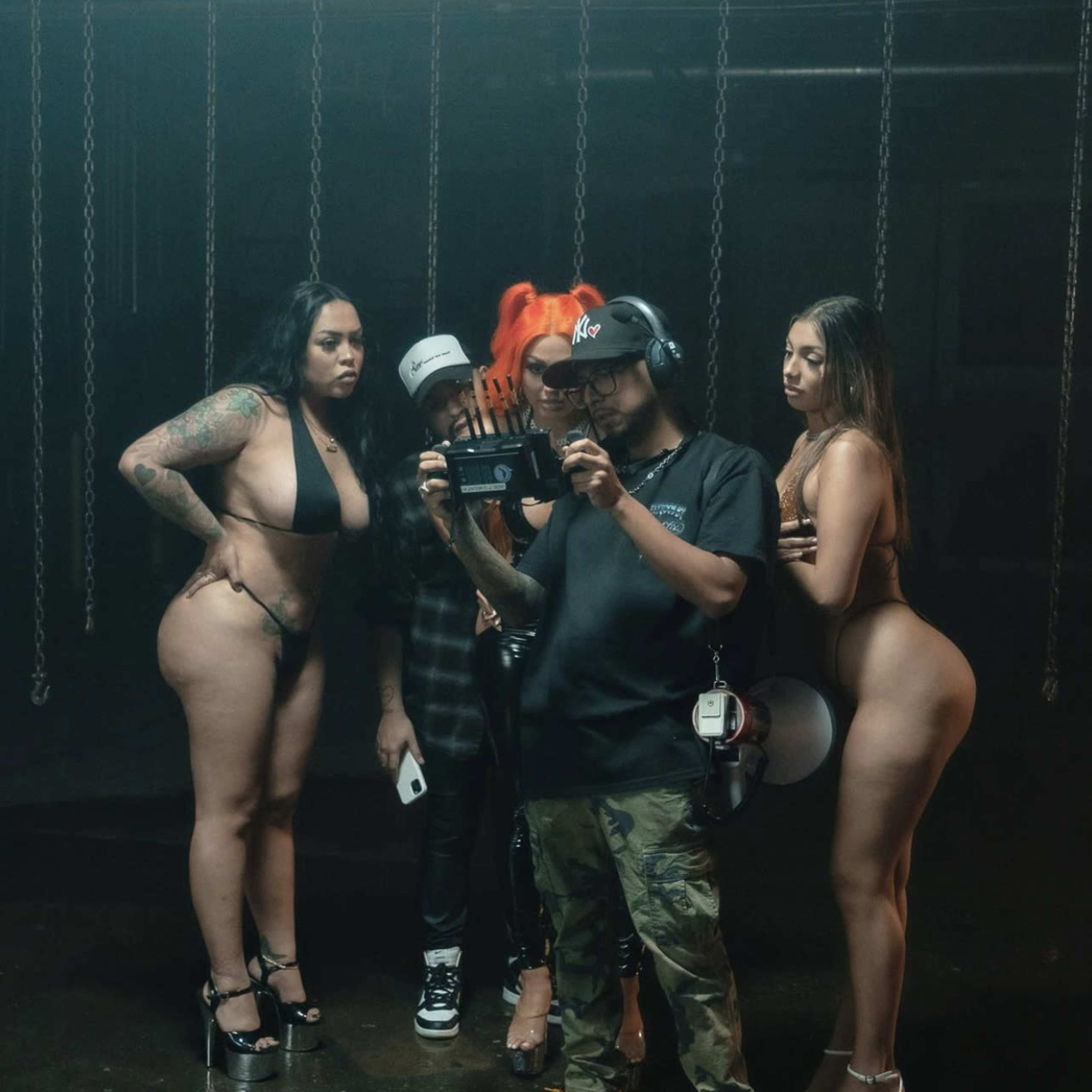 A group of five people, including three women in revealing outfits and a man holding a camera, are gathered in a dimly lit space with chains in the background.