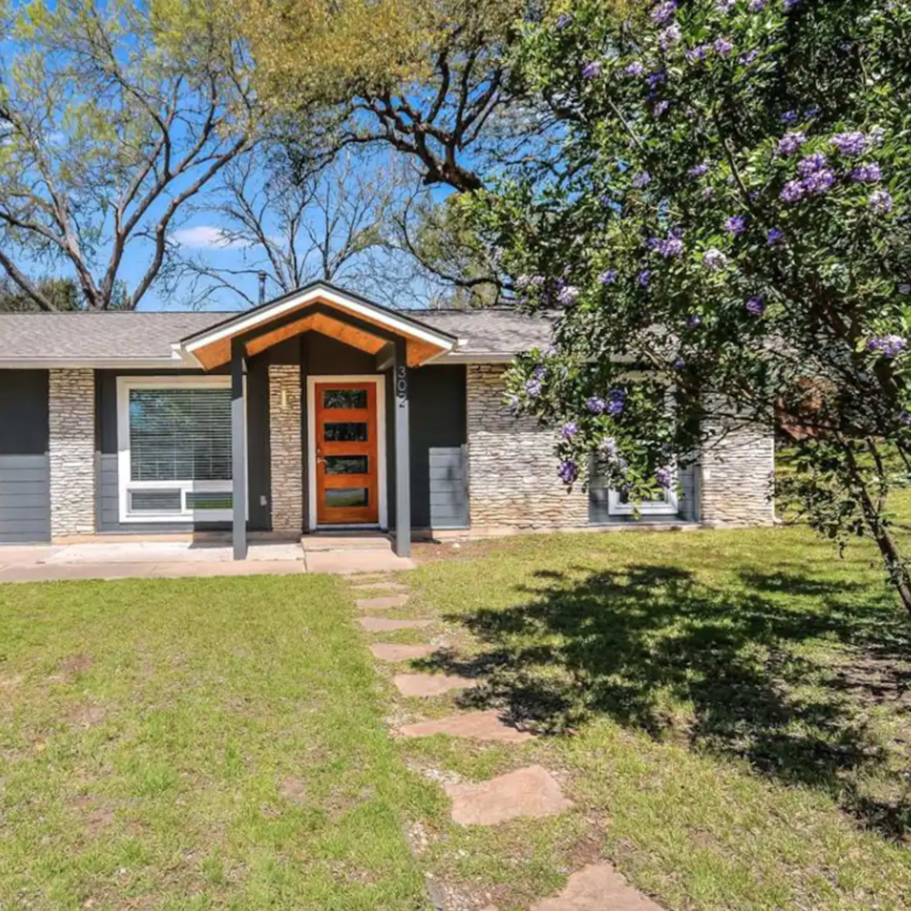 A single-story house with a stone and wood facade, featuring a front porch and a pathway lined with grass and stepping stones.