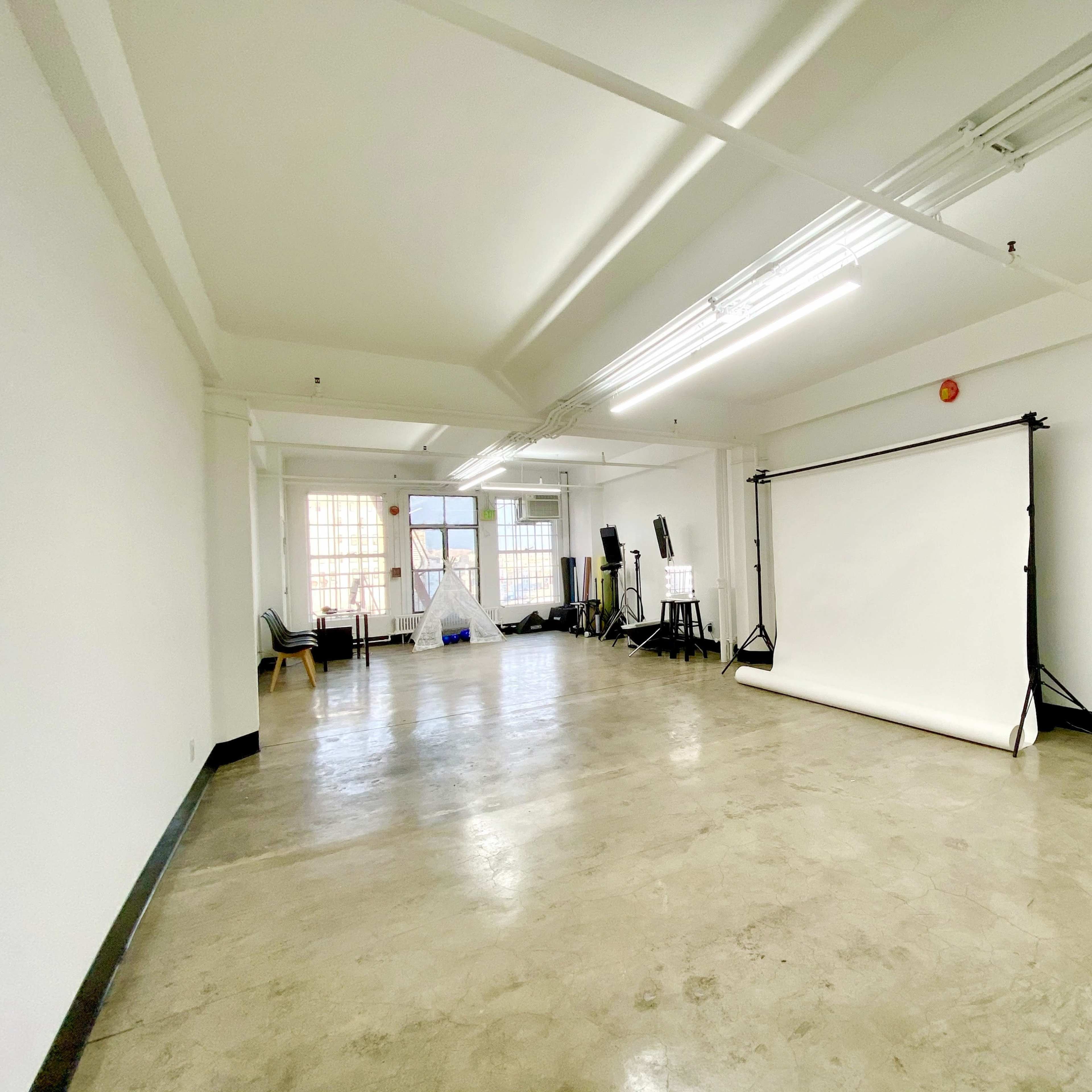 A spacious empty studio with concrete floors, white walls, and a backdrop setup at one end.