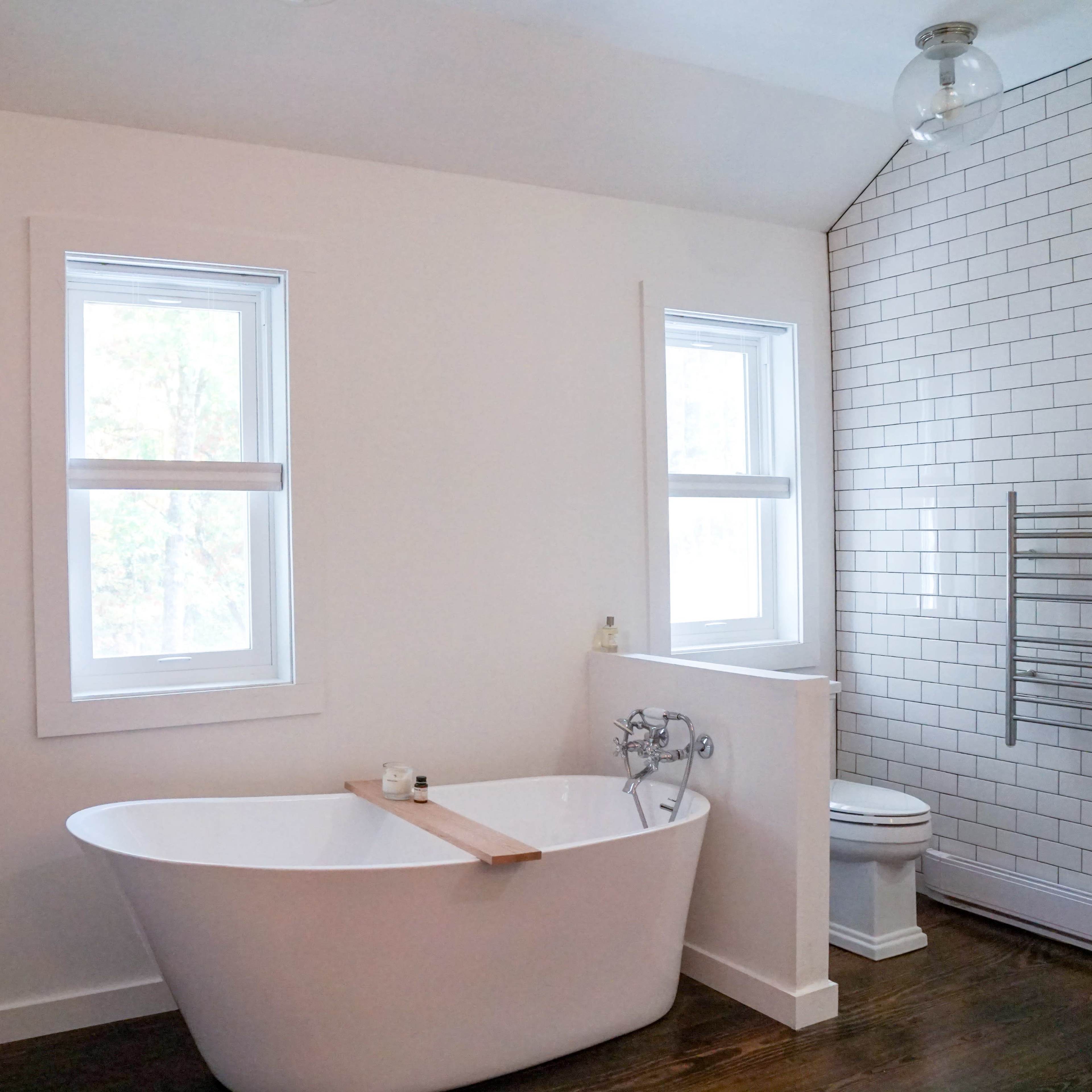 A modern bathroom featuring a freestanding white bathtub, a shower with white tiled walls, and a toilet, all bathed in natural light from two windows.