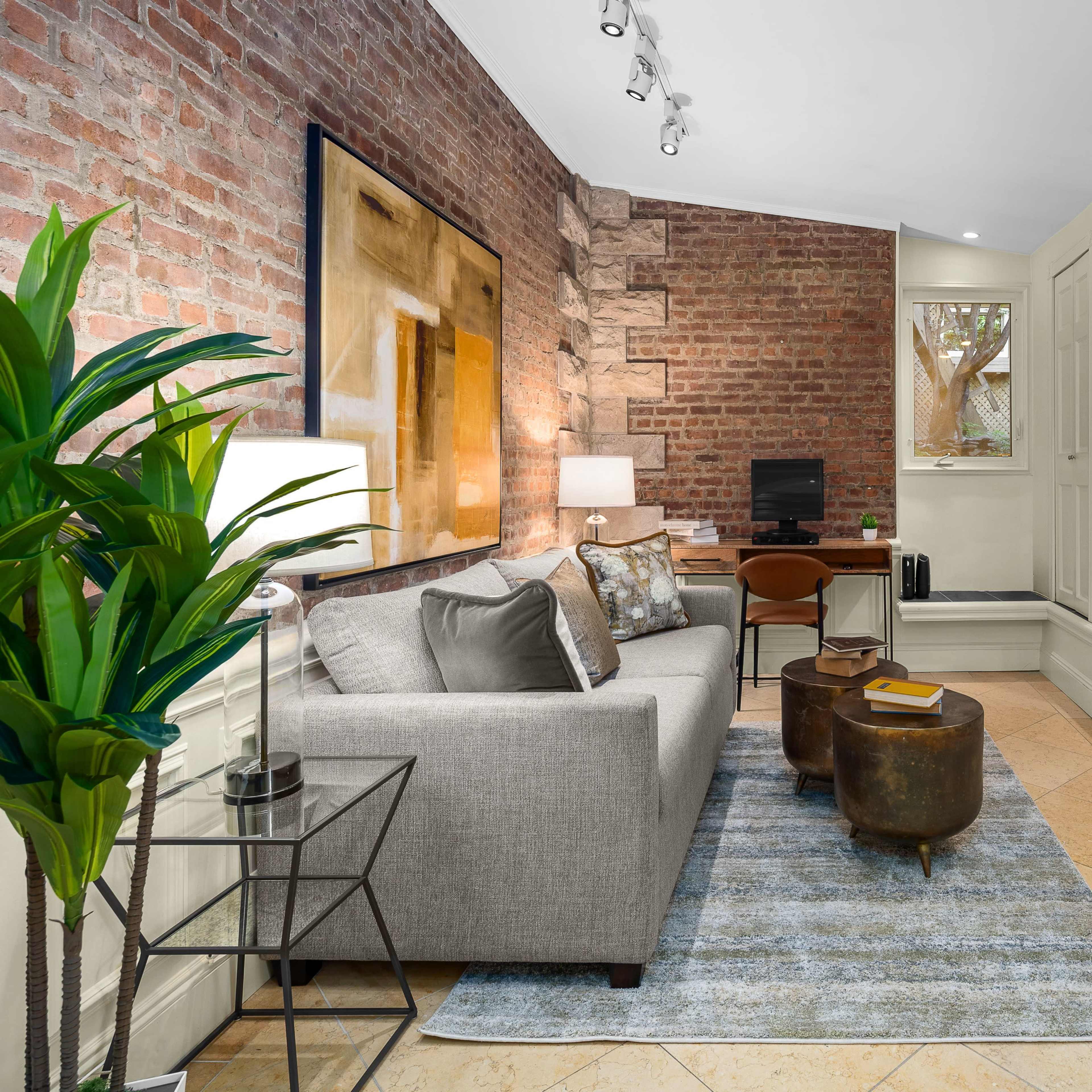 A cozy living room with a gray sofa, two metallic coffee tables, and a large window overlooking a small outdoor space, all set against a backdrop of exposed brick walls.