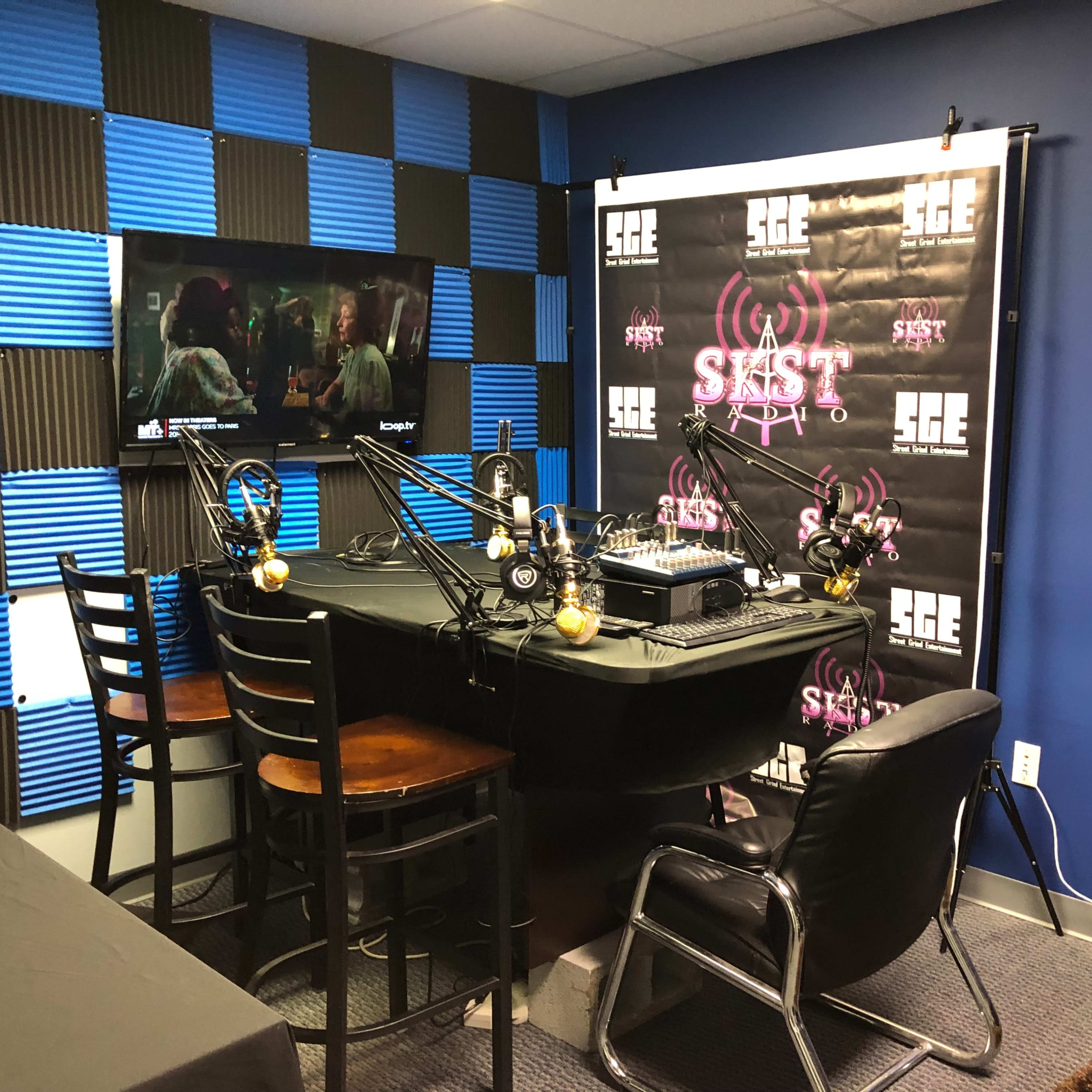 A recording studio with a black table, microphones, and chairs arranged around it, along with a television displaying a scene on the wall behind a backdrop featuring a logo.