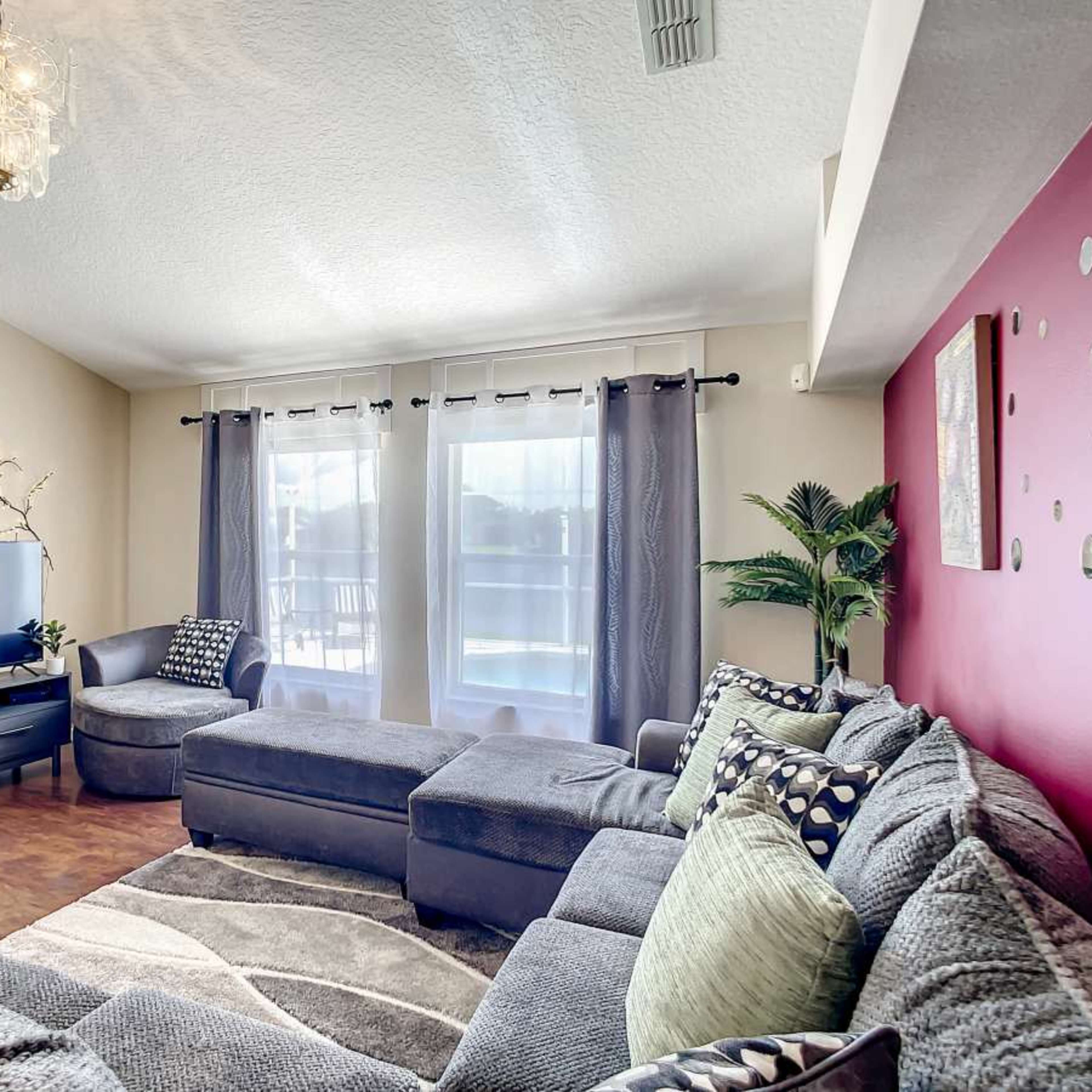 A cozy living room with a sectional sofa, a television, and large windows covered by sheer curtains, complemented by potted plants and decorative wall elements.