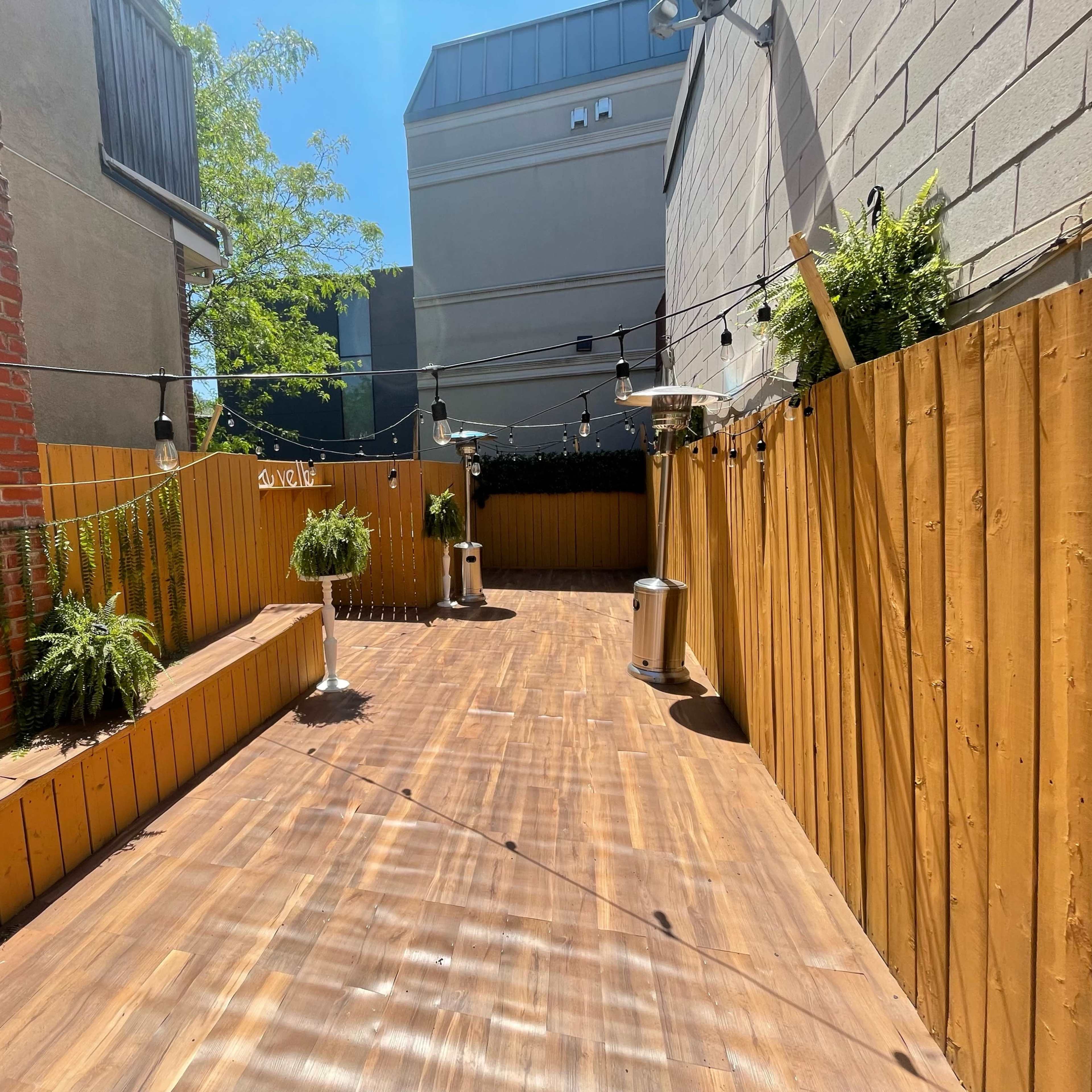 A narrow, sunlit outdoor patio features wooden decking, enclosed by wooden fencing and adorned with potted plants and hanging lights.