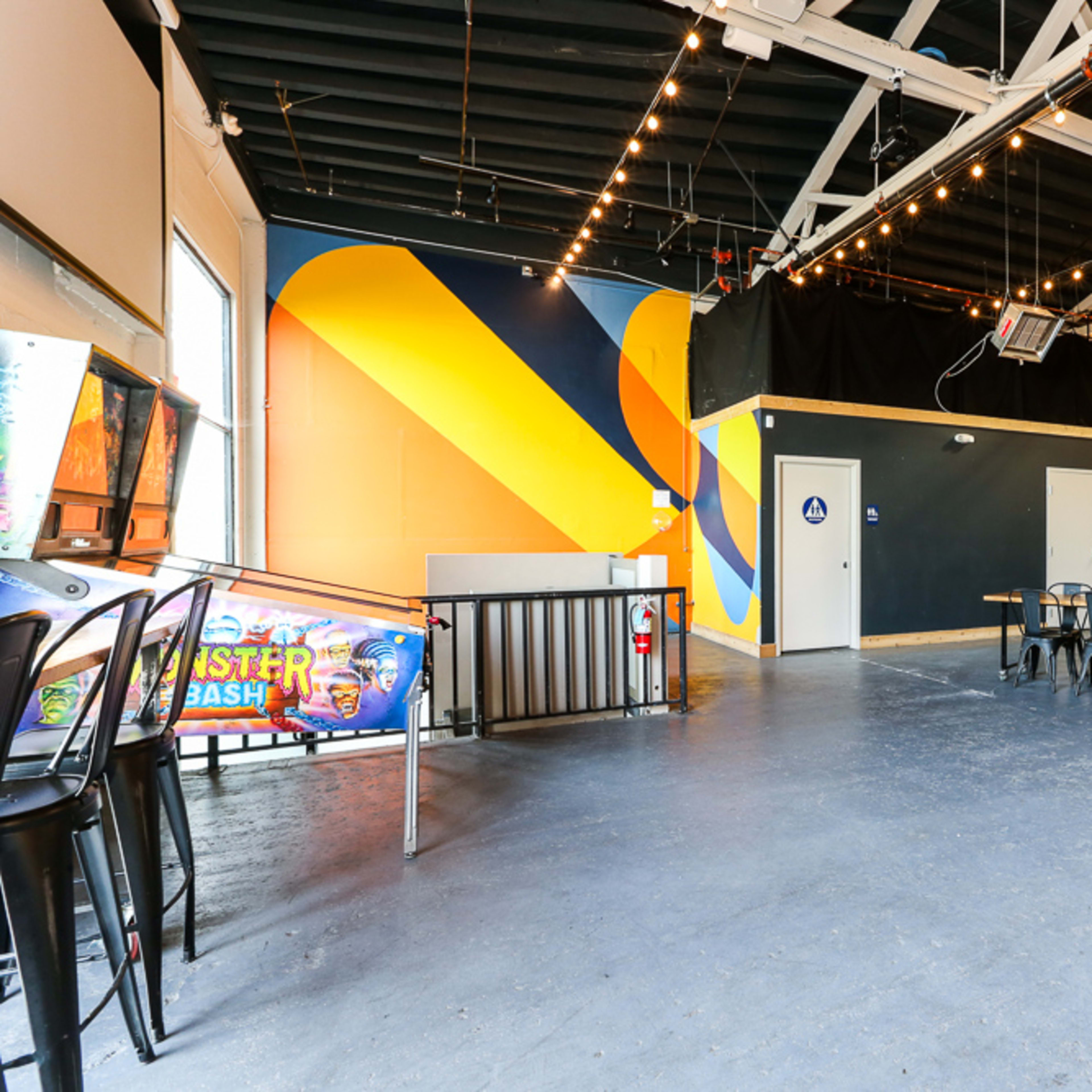 A spacious interior of a venue featuring colorful wall art, seating arrangements of metal chairs and tables, and a bar area.
