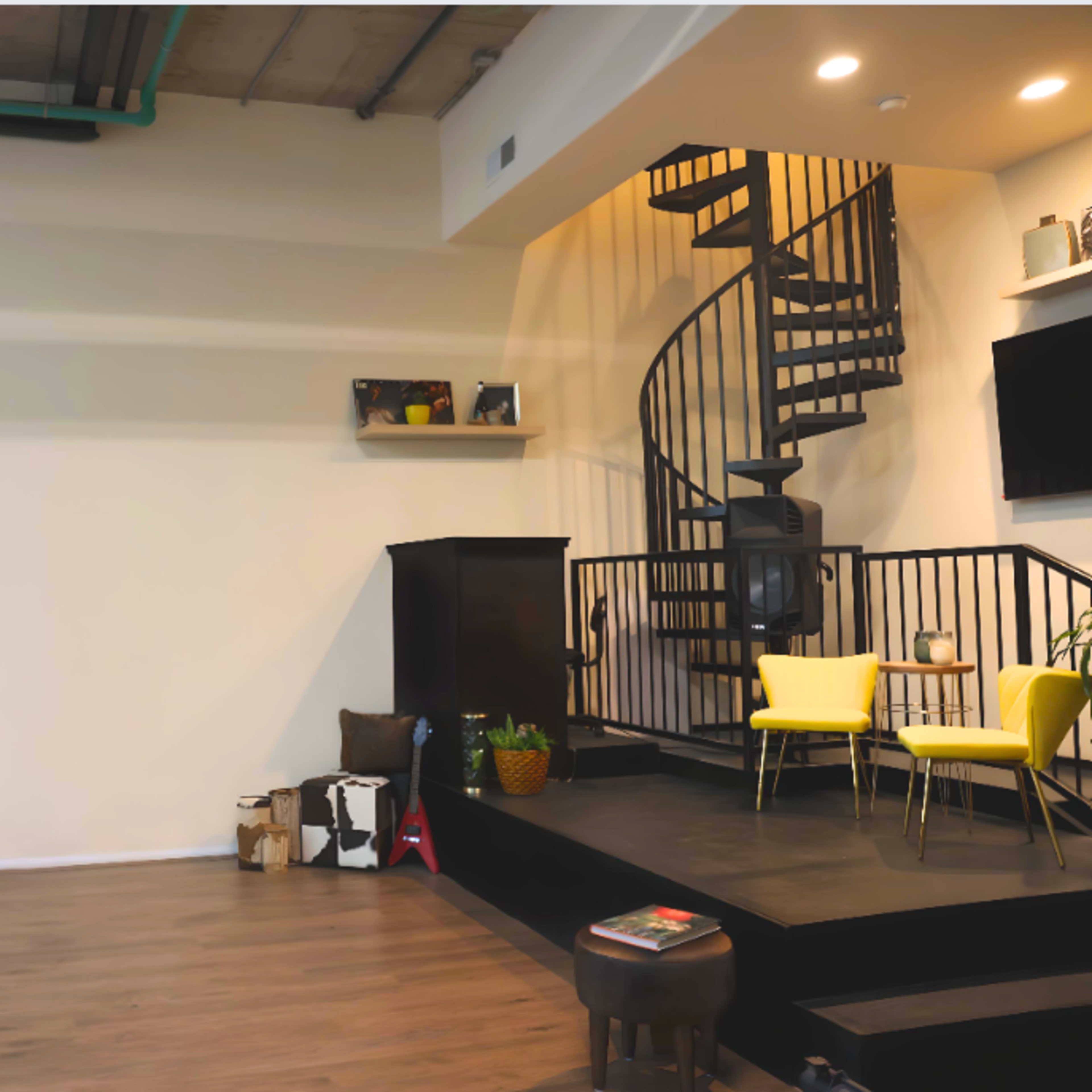 A modern interior space featuring a spiral staircase, a black platform, yellow chairs, and a cozy seating area with a lamp.