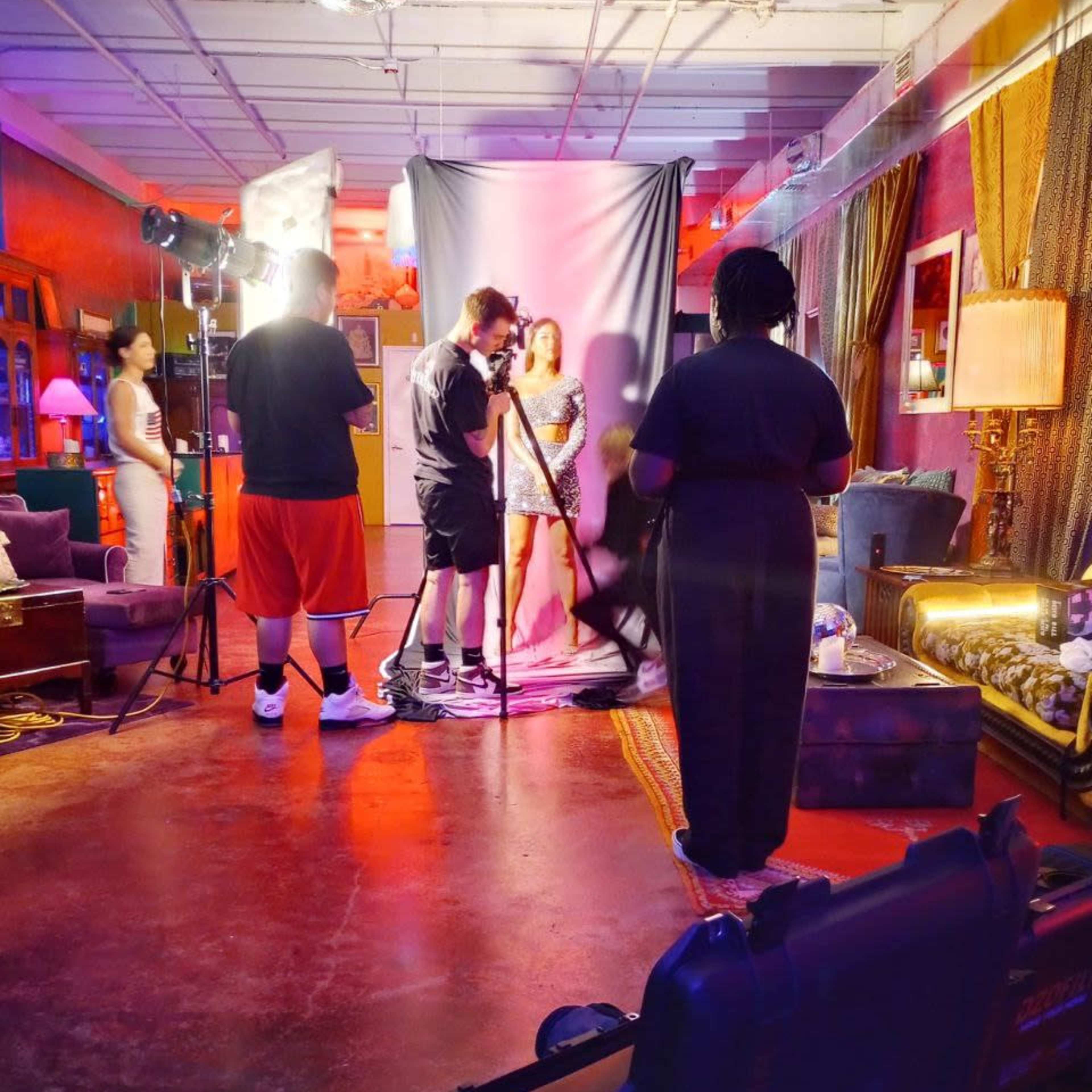 A film crew sets up a photoshoot in a colorful studio, with a model posing in front of a backdrop while crew members adjust lighting and equipment.
