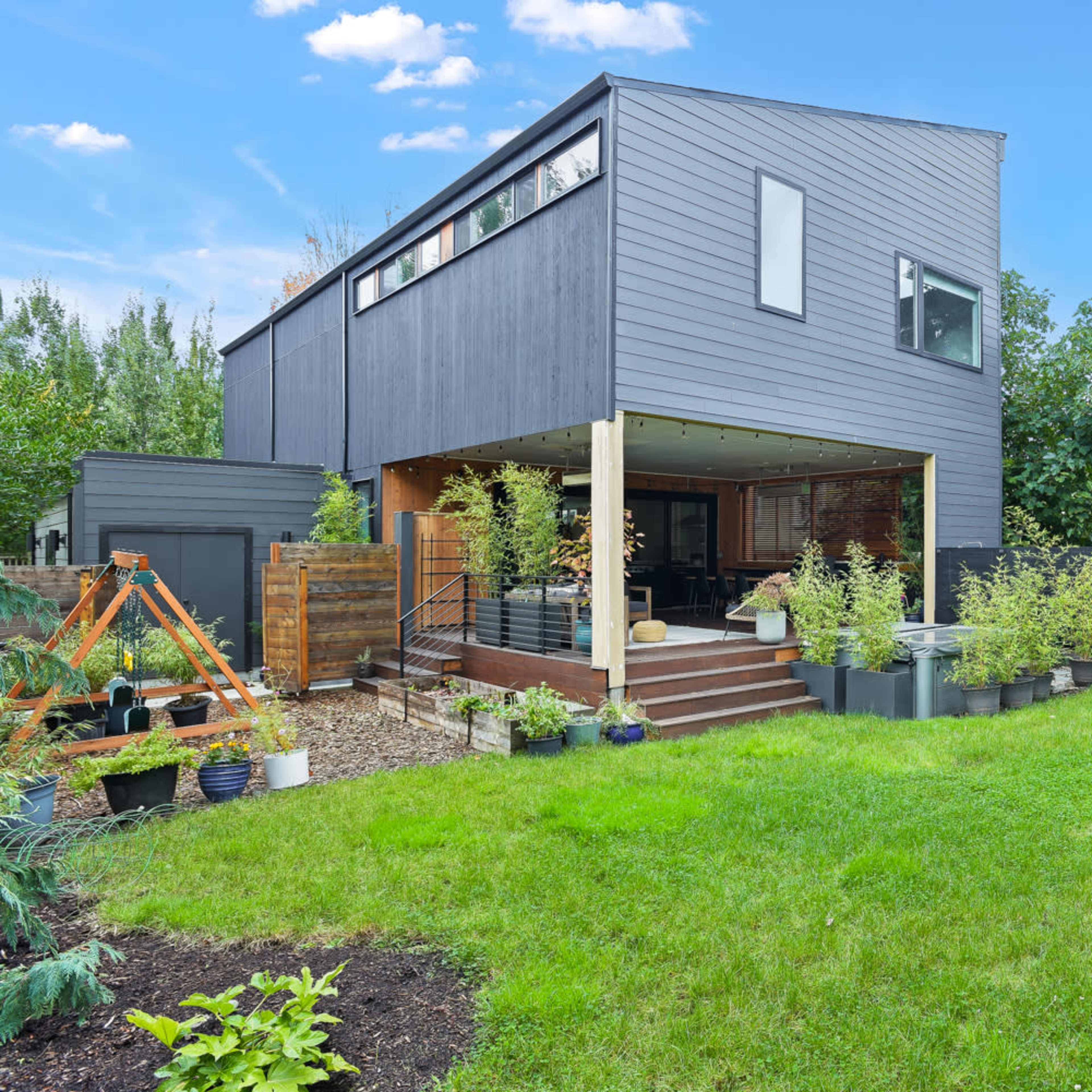 A modern two-story house with a wooden deck and landscaped yard is surrounded by greenery and garden planters.