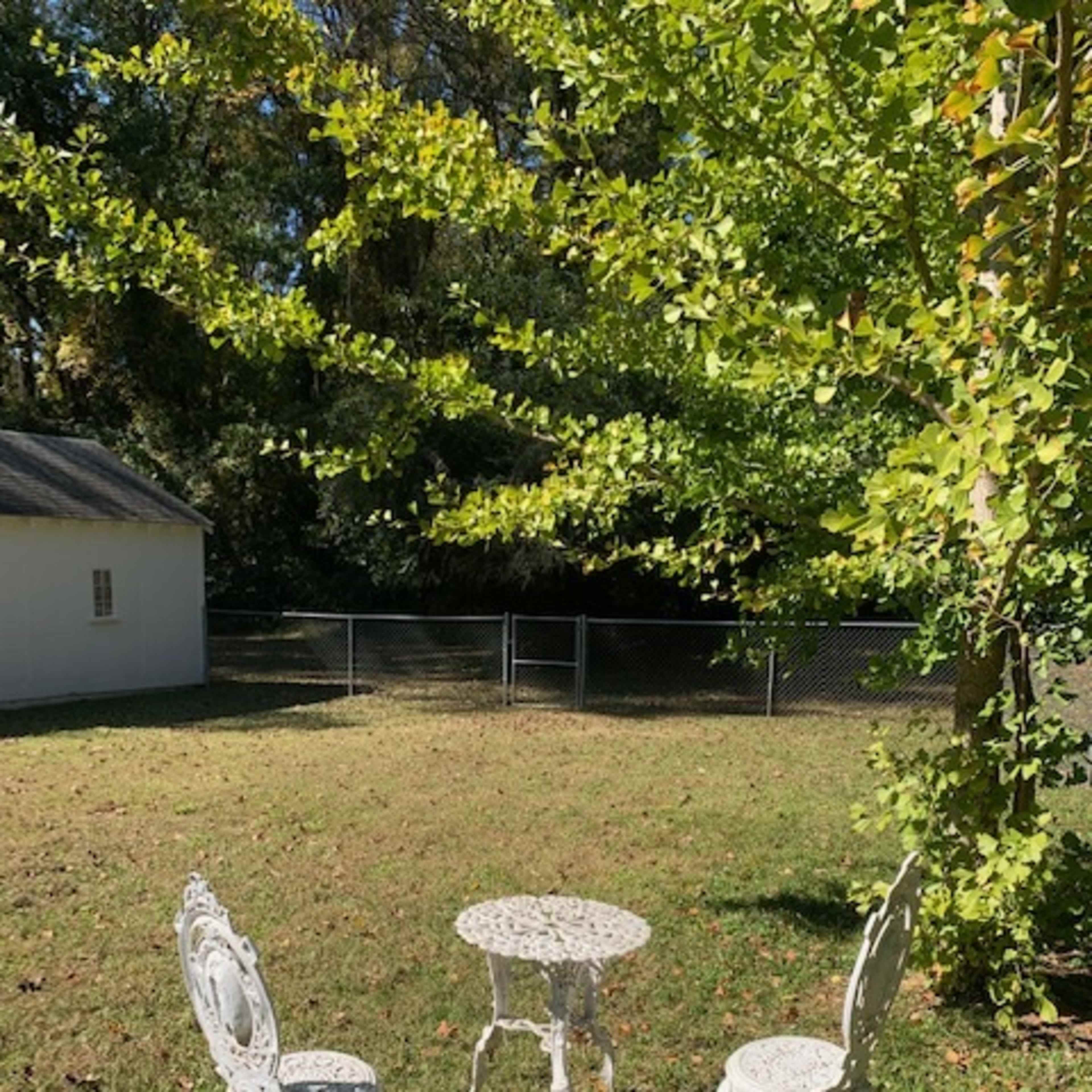A small white metal table with three matching chairs is placed on a grassy area surrounded by trees and a fence.