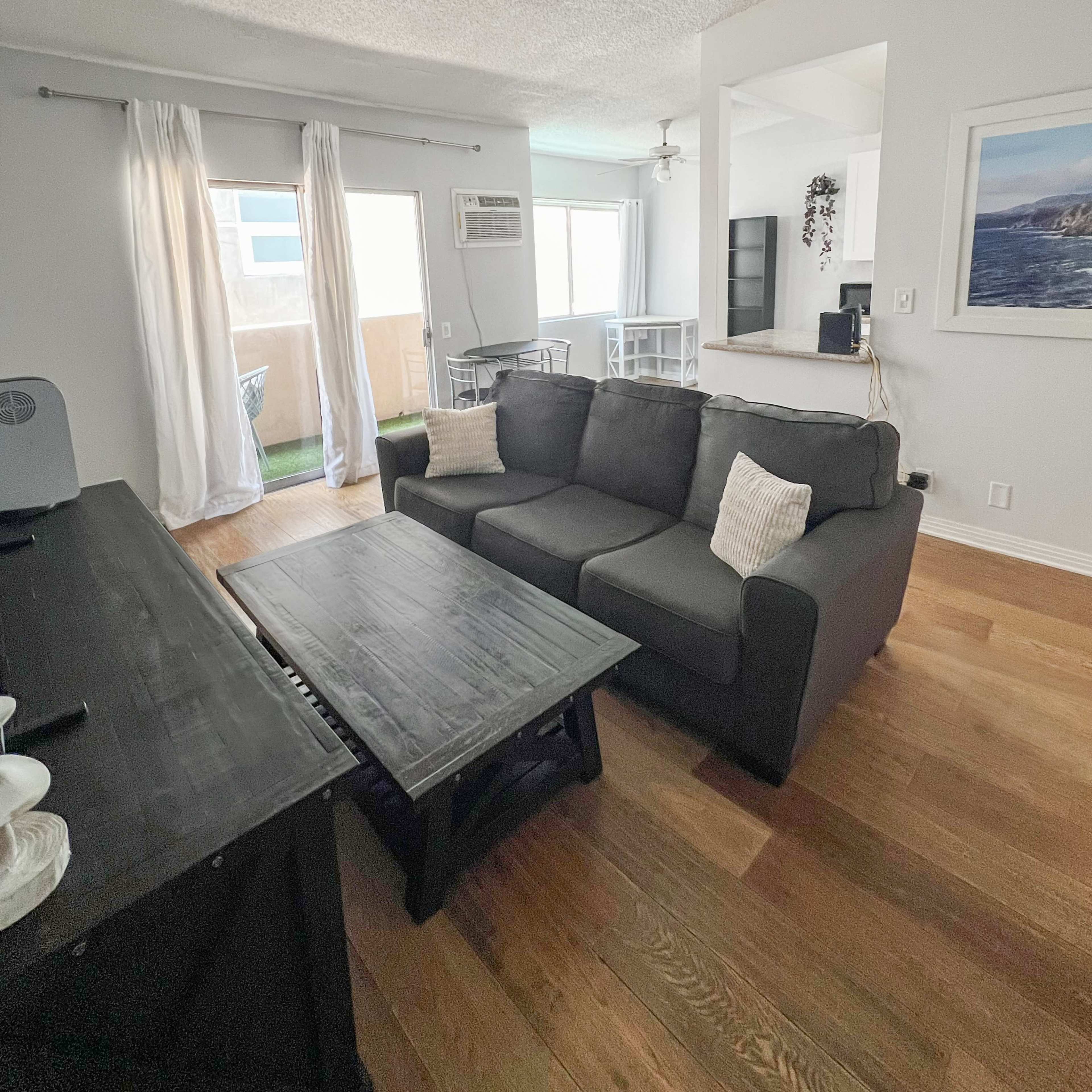 A living room with a dark gray couch, a wooden coffee table, and large windows with white curtains, leading to a balcony.