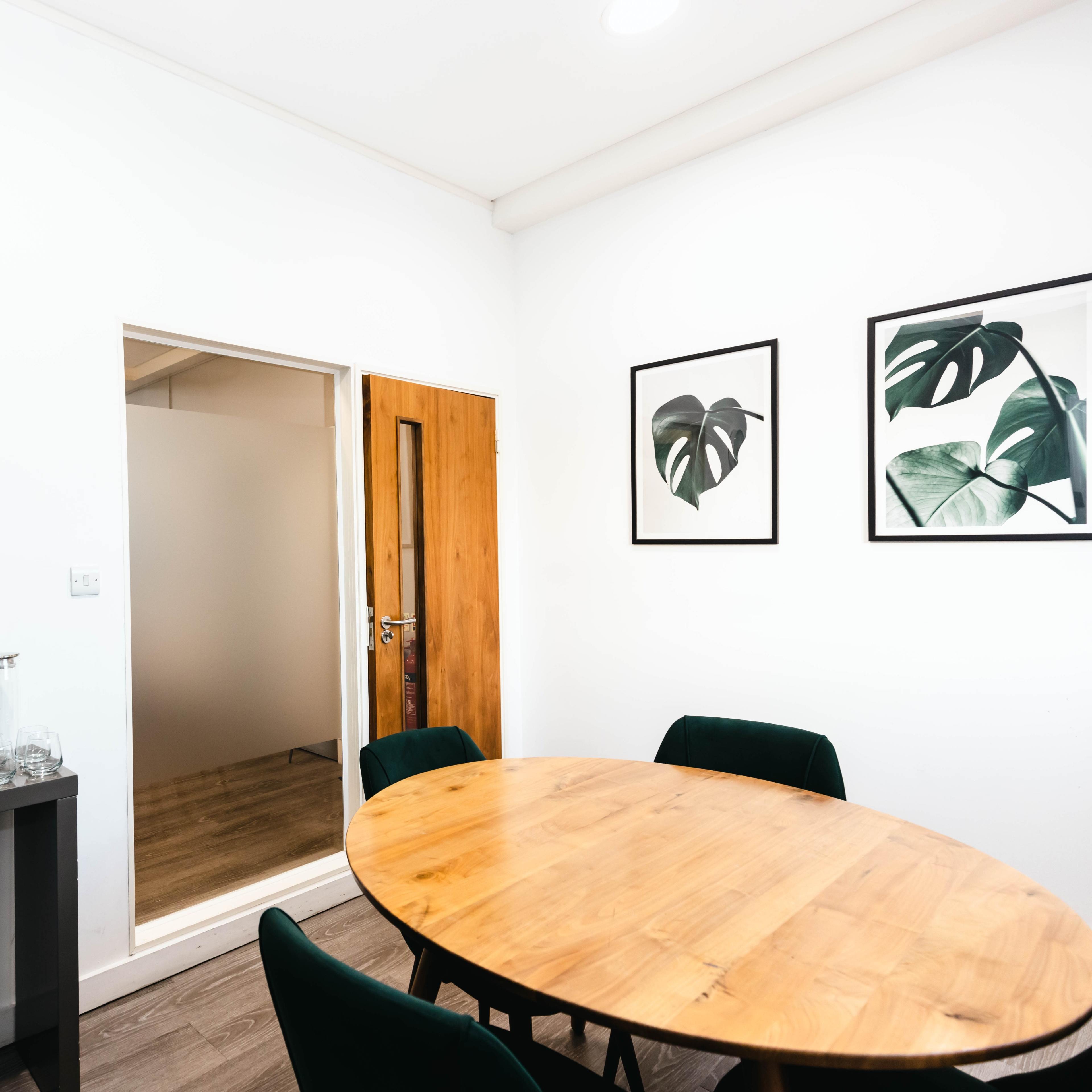 A round wooden table is surrounded by dark green chairs in a room with a clock, a framed plant print, and a door leading to another space.