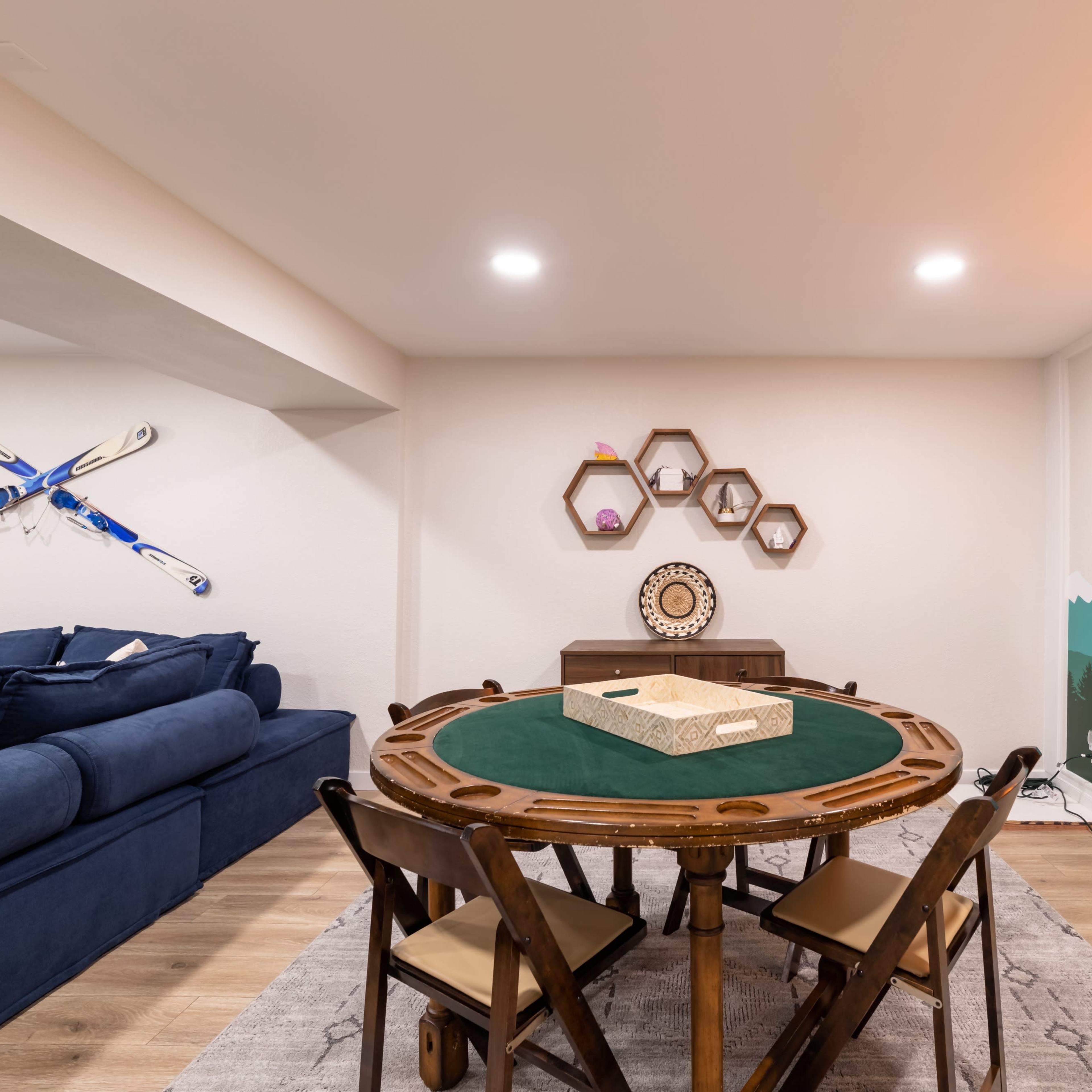 A cozy basement game area featuring a round table with a green felt surface, surrounded by wooden chairs, and a decorated wall with mountain mural art and a neon sign.