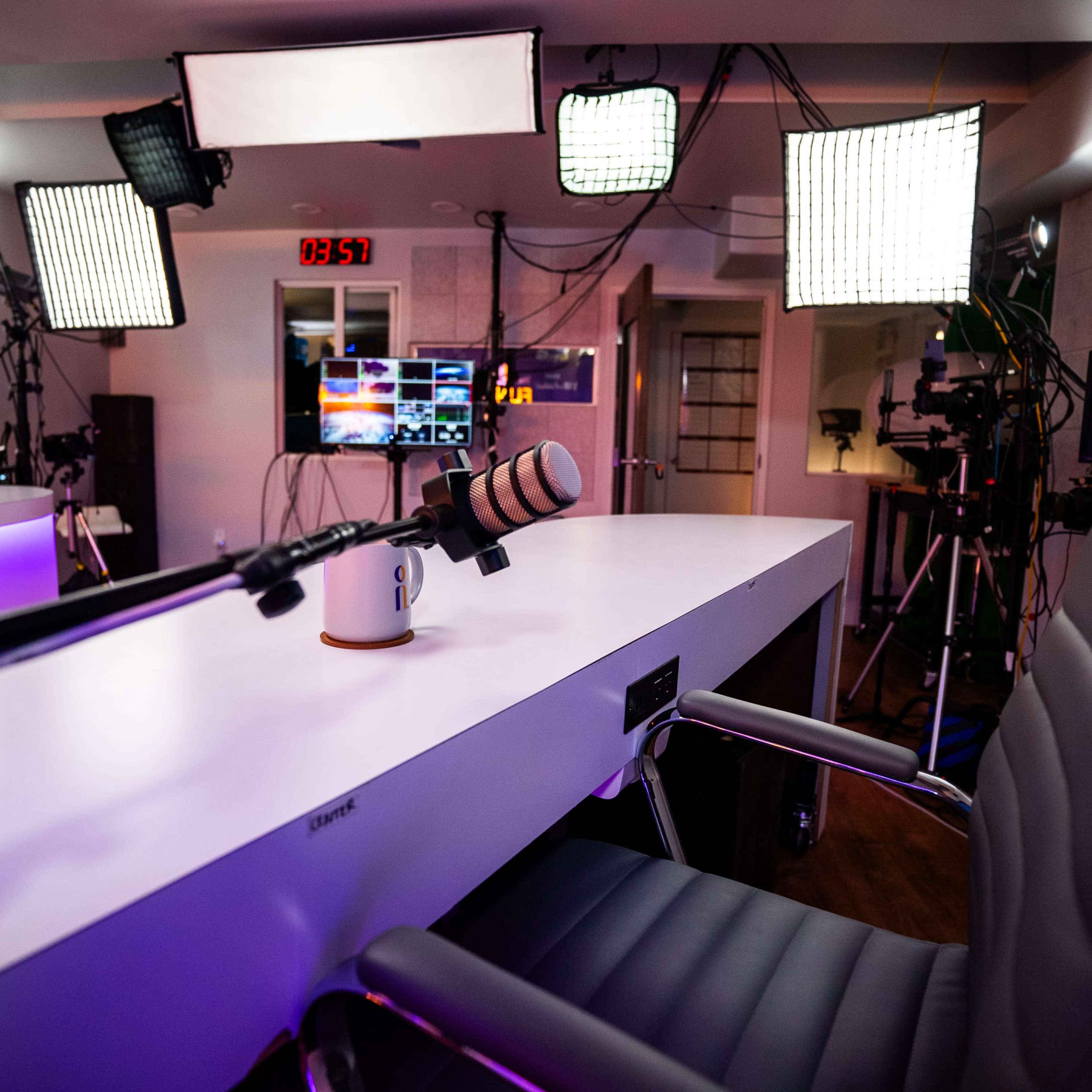 A recording studio setup features a large table with a microphone, an office chair, and multiple lights positioned around the space.