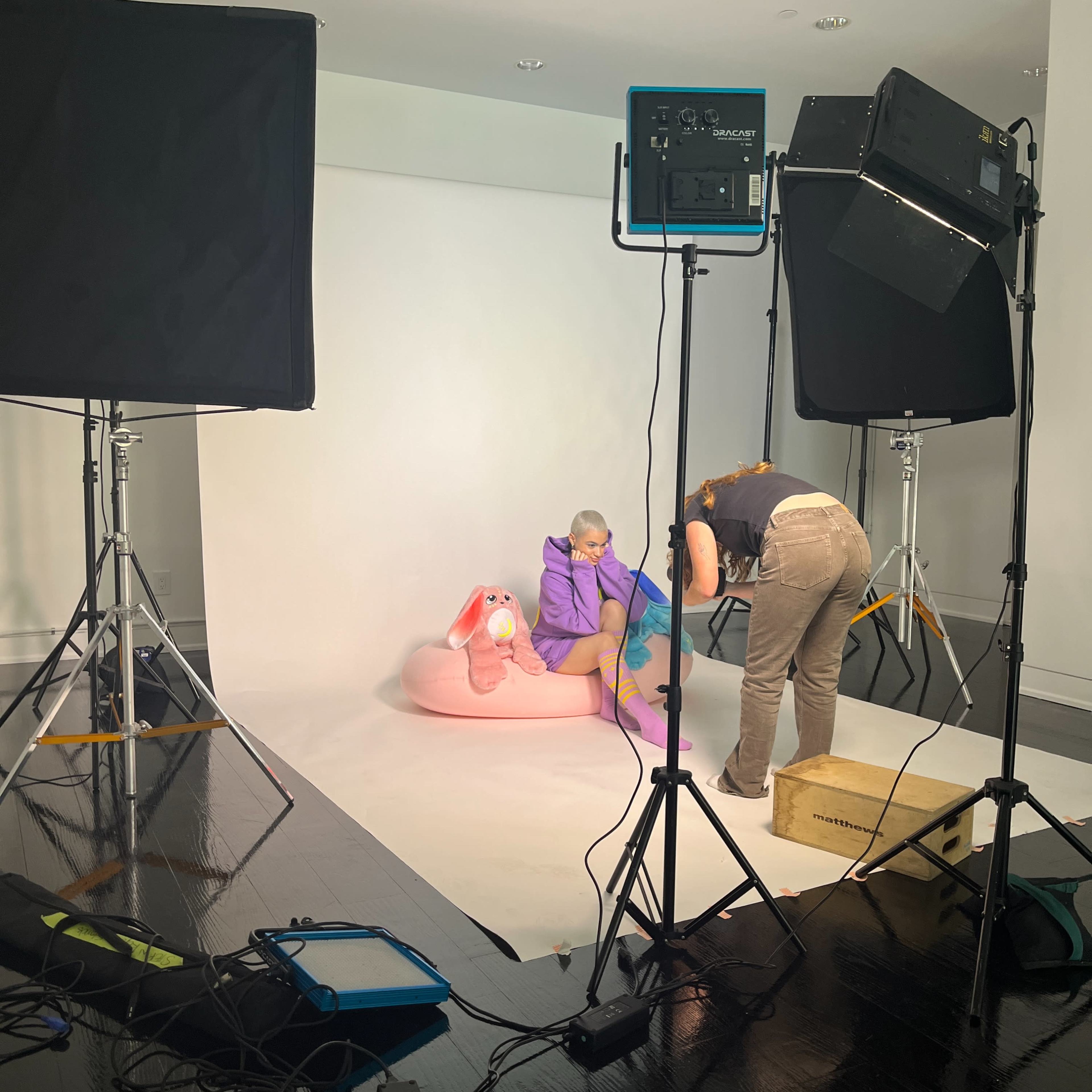 A photo shoot is taking place in a studio with lighting equipment, where a person sits on a pink bean bag next to a large stuffed rabbit while another person adjusts a prop.