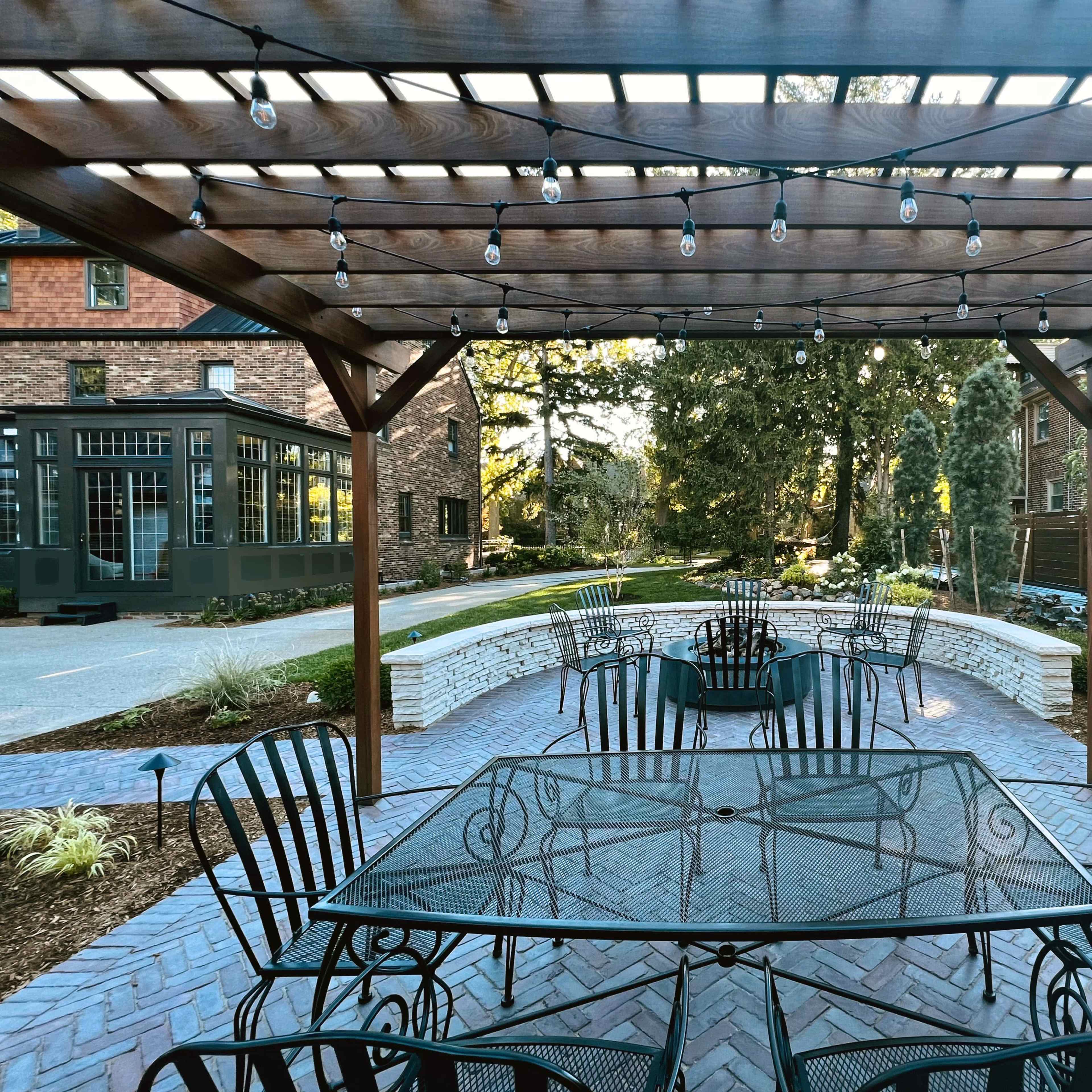 A patio area with several black metal tables and chairs is covered by a wooden pergola adorned with string lights, overlooking a landscaped backyard.