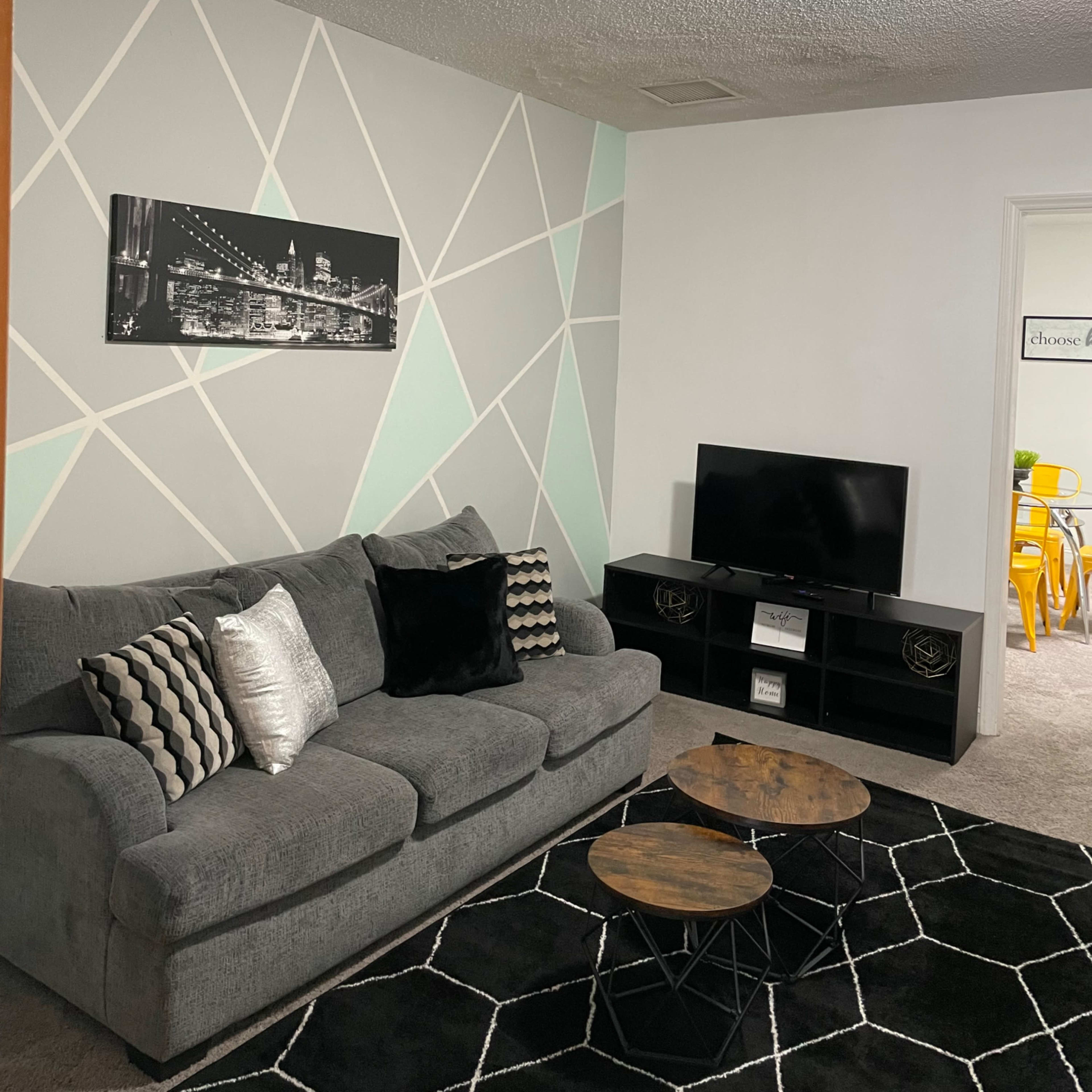 A living room featuring a gray sofa with decorative pillows, two wooden coffee tables, a black television stand, and a geometric patterned accent wall.