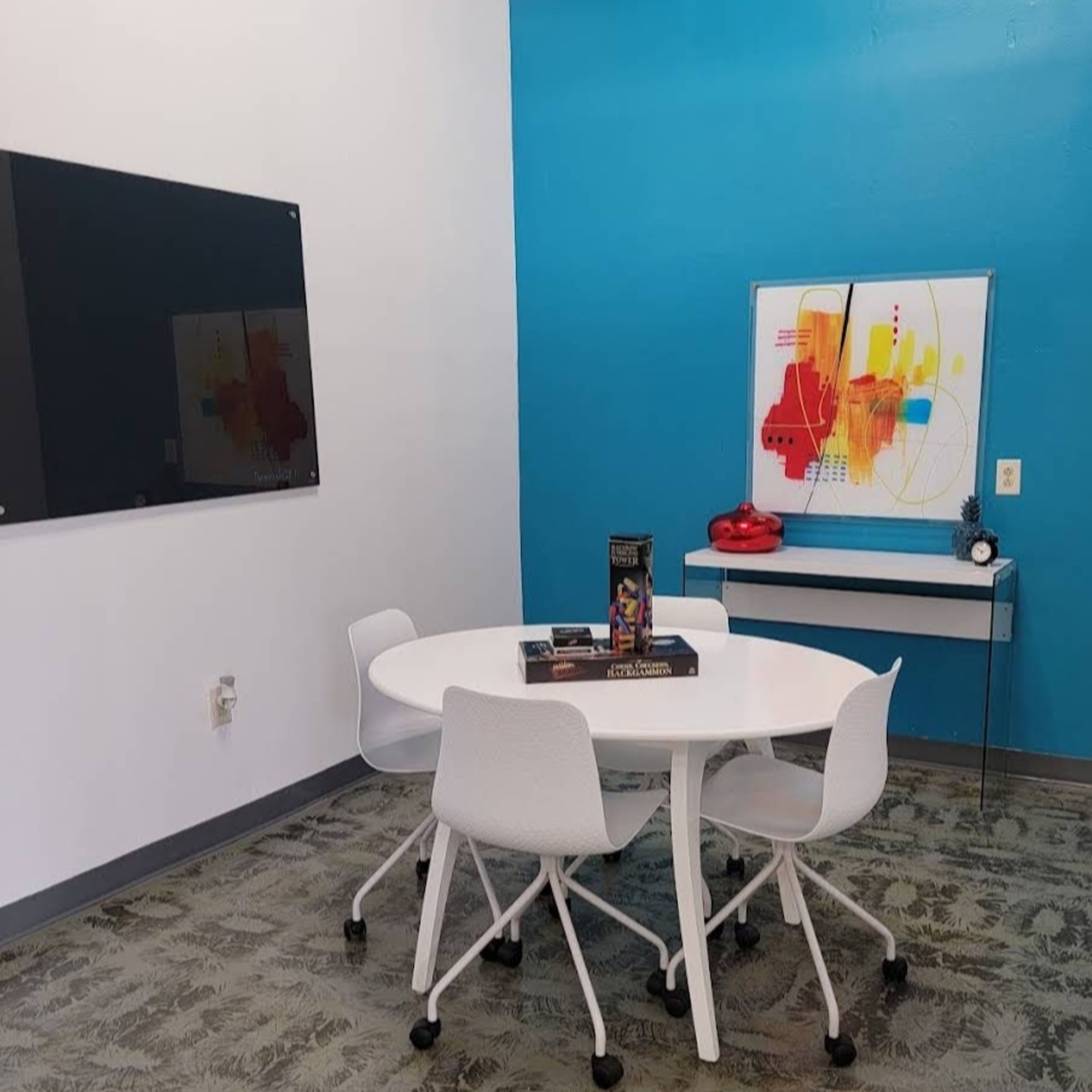 A small conference room features a round table surrounded by four white chairs, a black wall-mounted screen, and a colorful abstract artwork on the wall.