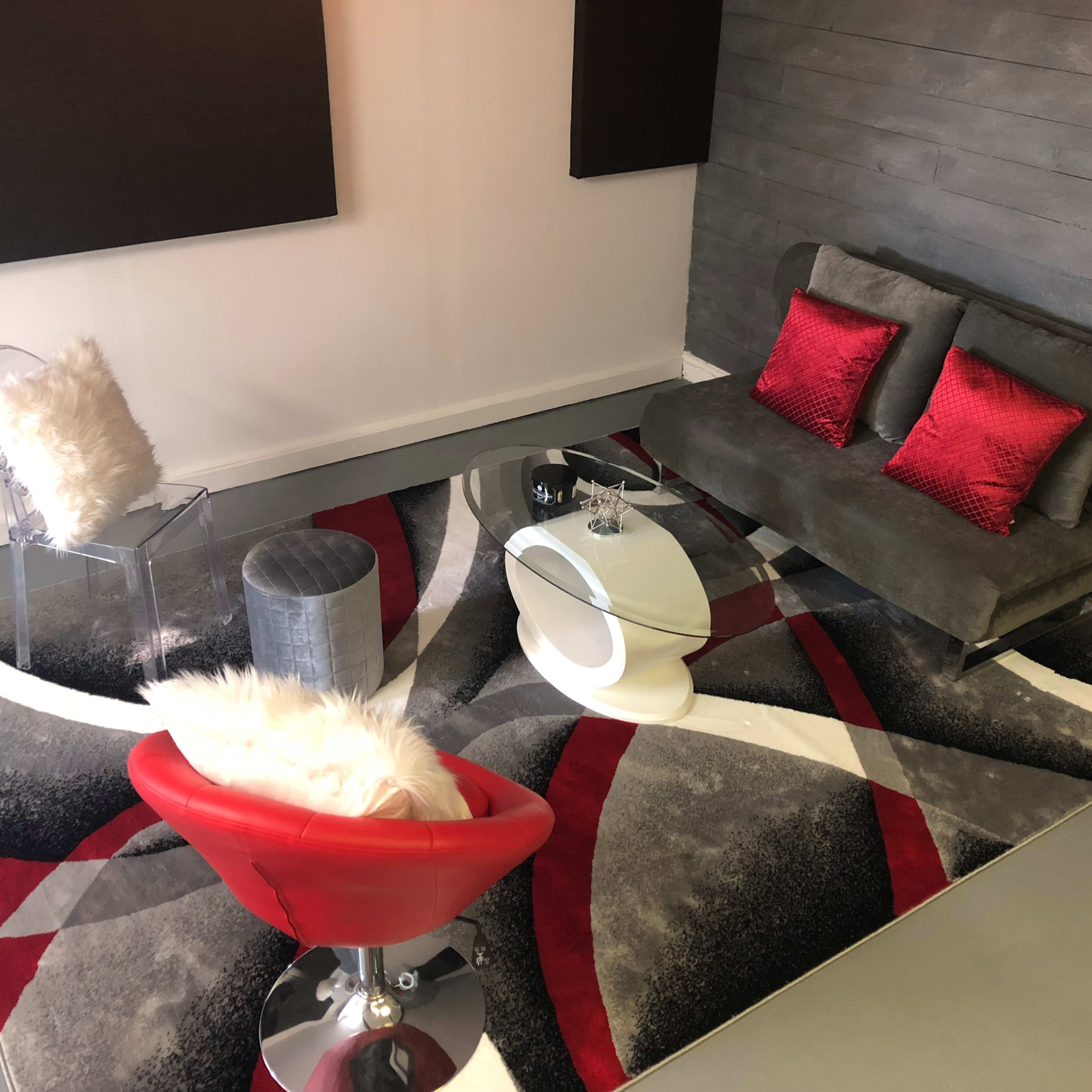 The scene features a modern lounge area with a gray sofa, red accent pillows, a glass coffee table, a silver stool, a clear chair with a white pillow, and a patterned area rug.