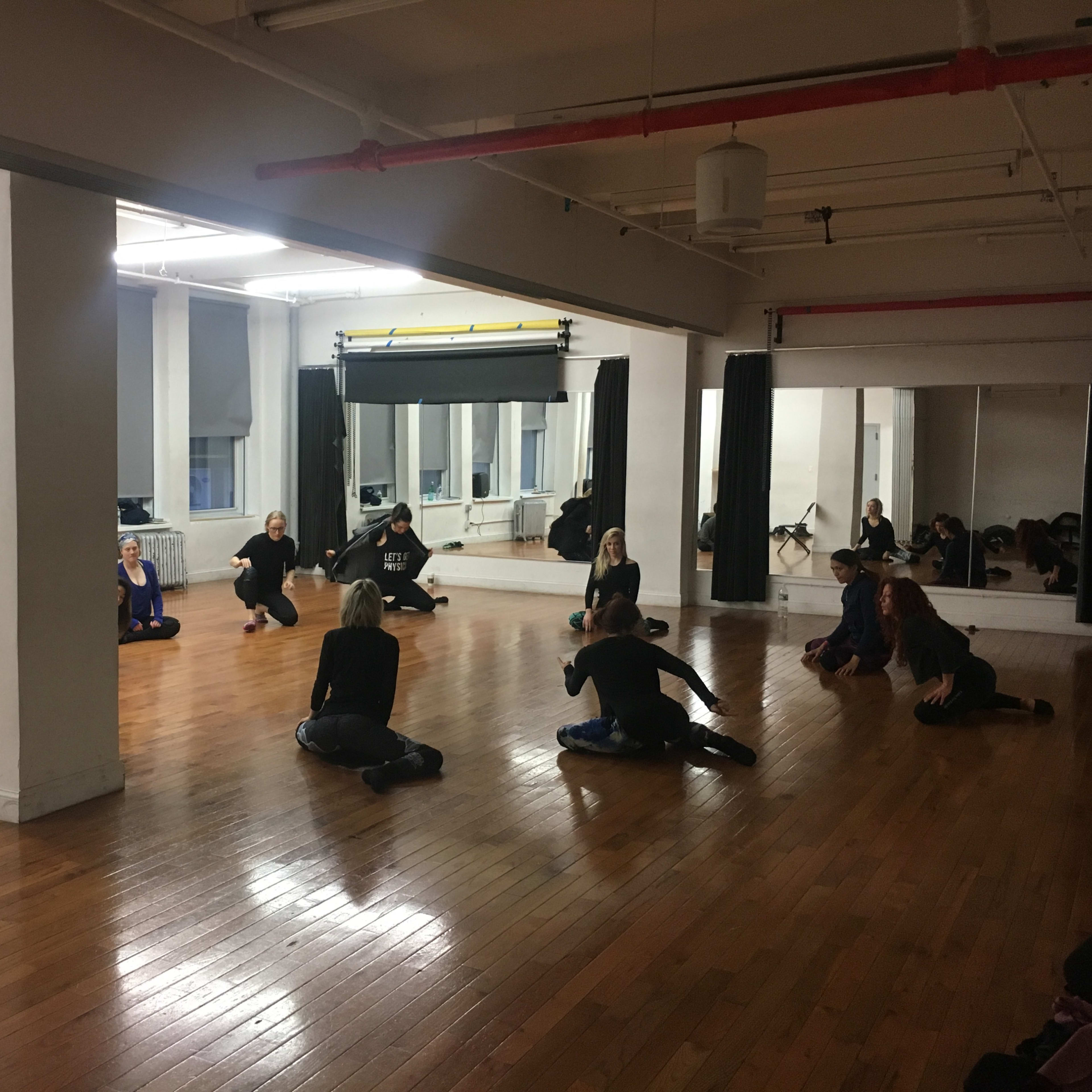 A group of individuals engages in a stretching exercise in a spacious dance studio with hardwood floors and mirrors.