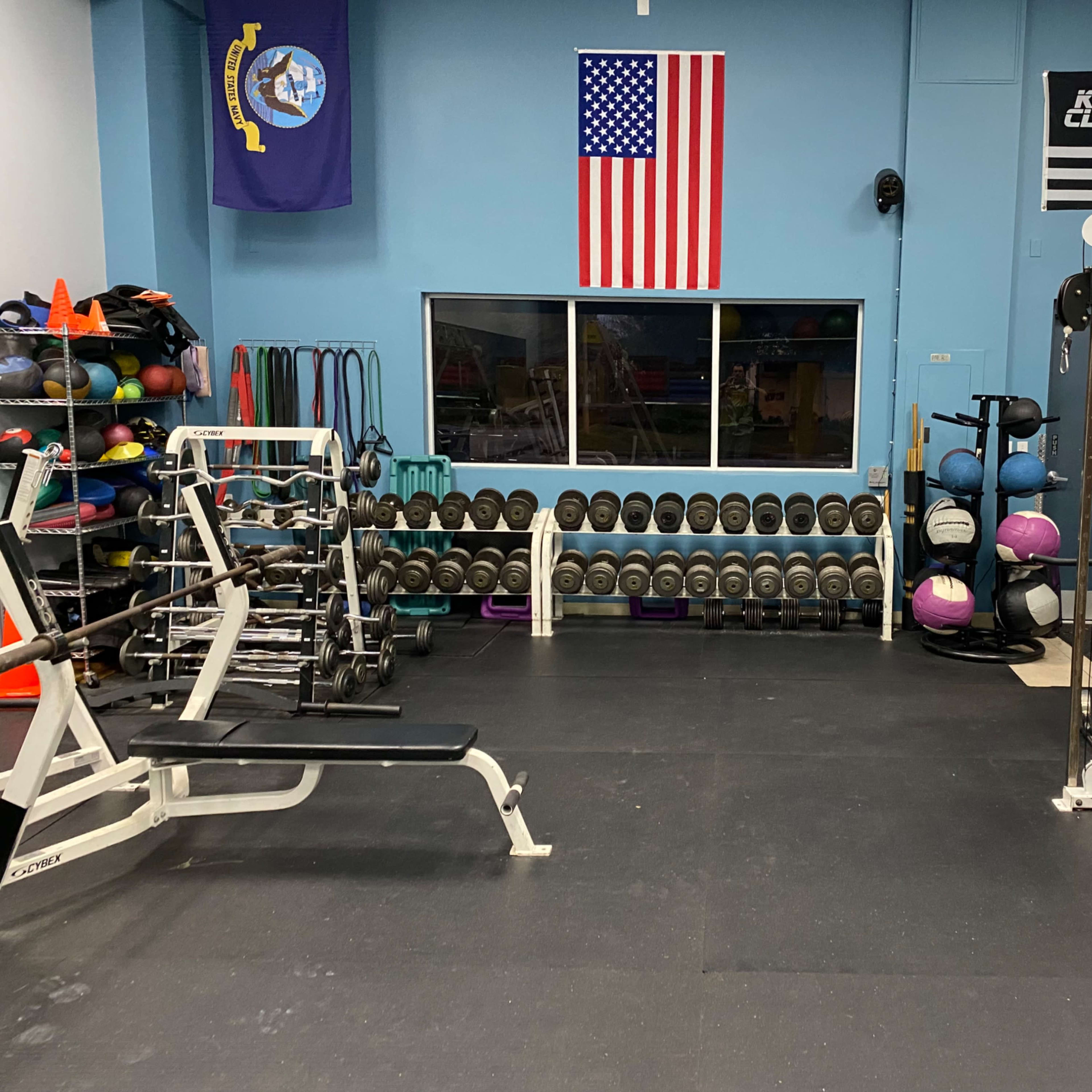 A gym interior with weightlifting equipment, exercise balls, and free weights lined up against the wall, adorned with American and military flags.