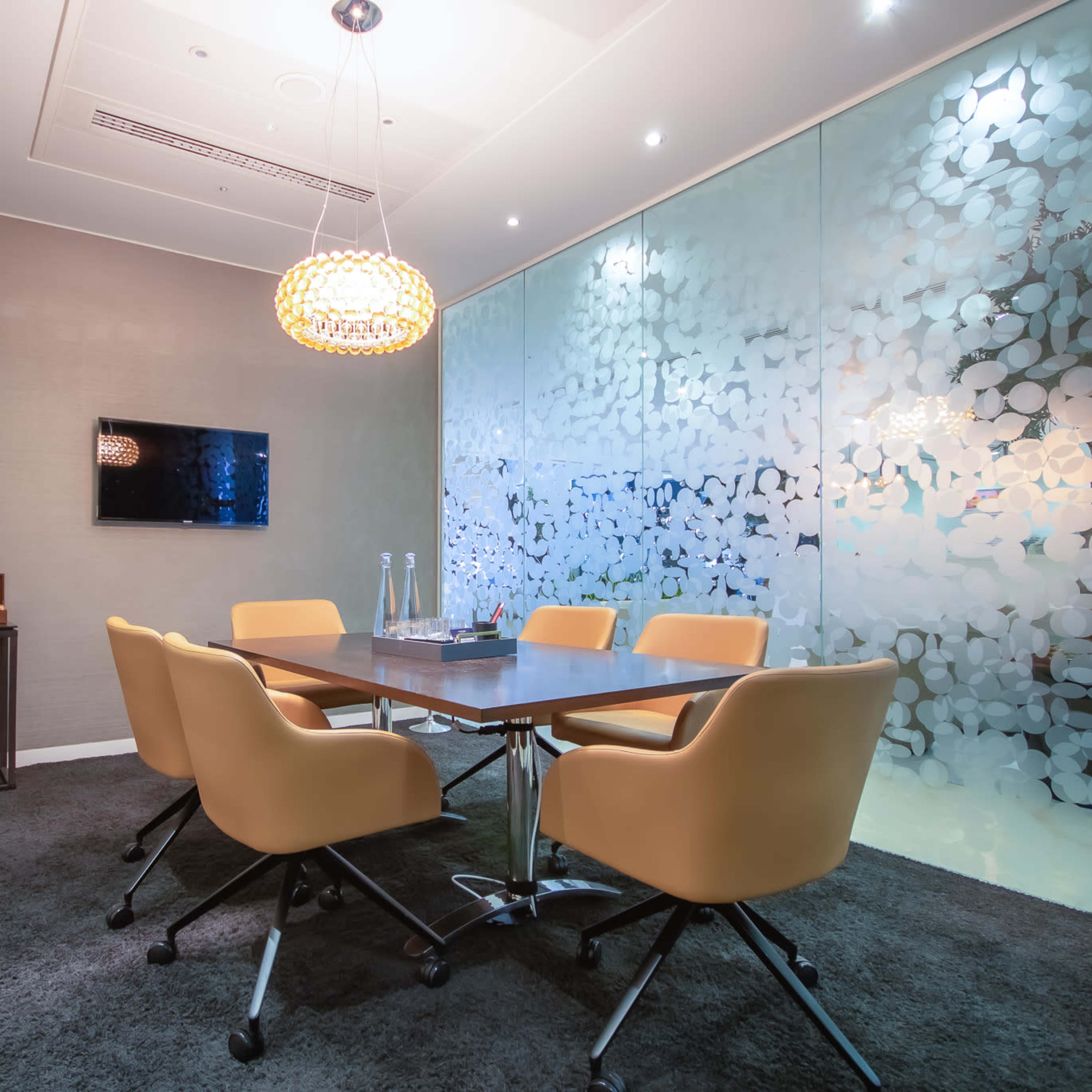 A modern meeting room features a large table surrounded by six orange chairs, with frosted glass walls and a wall-mounted television.