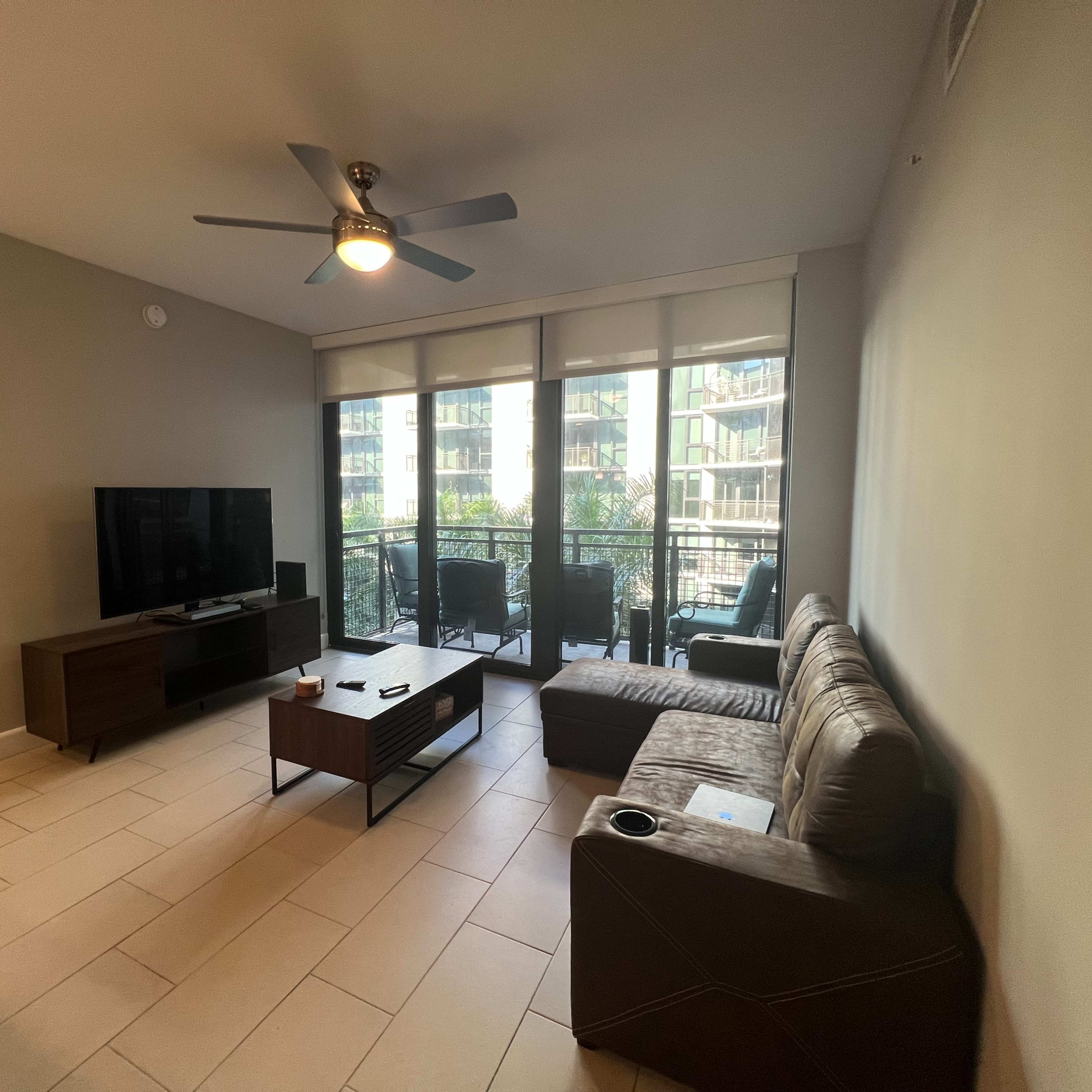 A living room with a brown leather sofa, a coffee table, a television on a stand, and large windows overlooking a balcony.