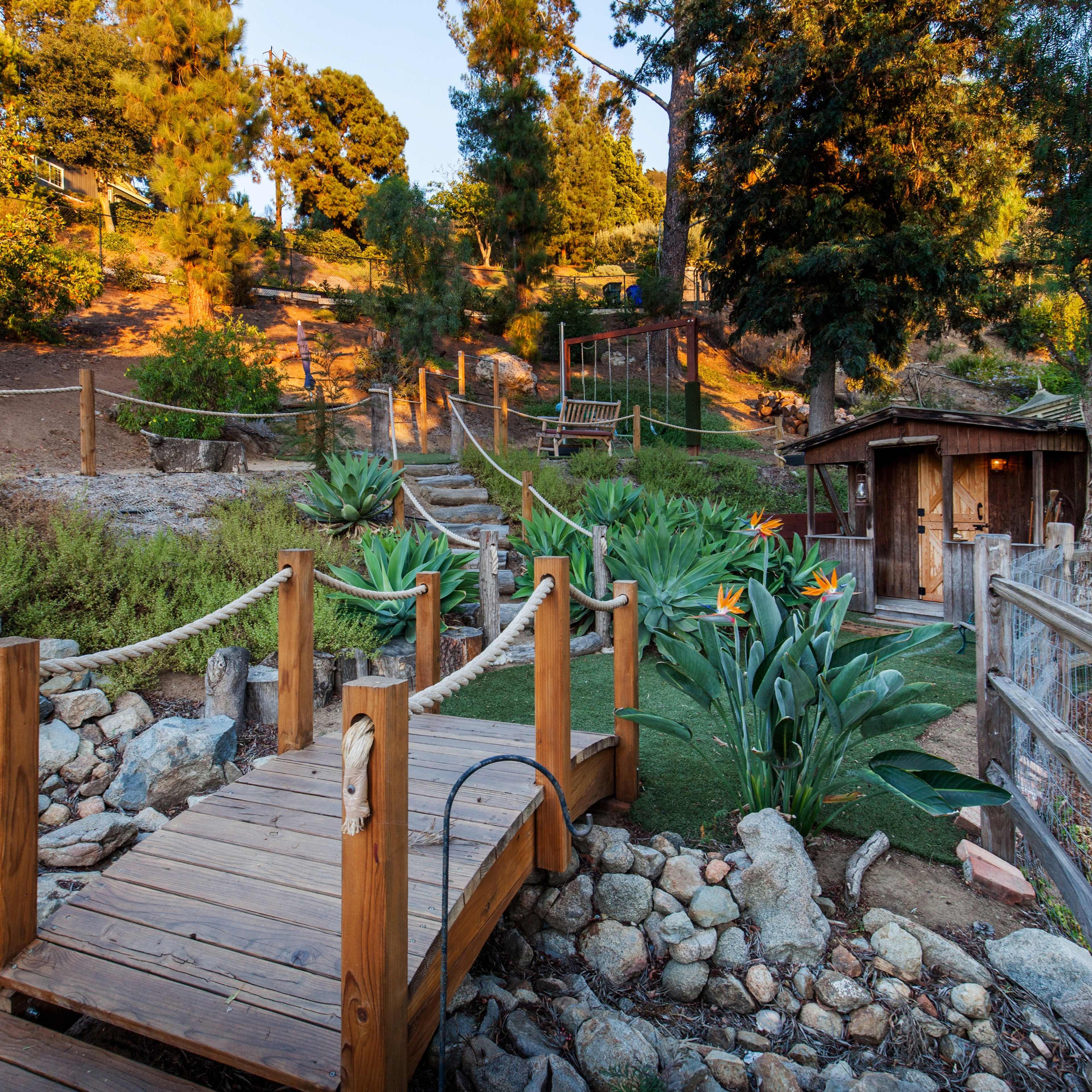 A wooden bridge connects a landscaped area with pathways leading through greenery and a rustic shed in the background.