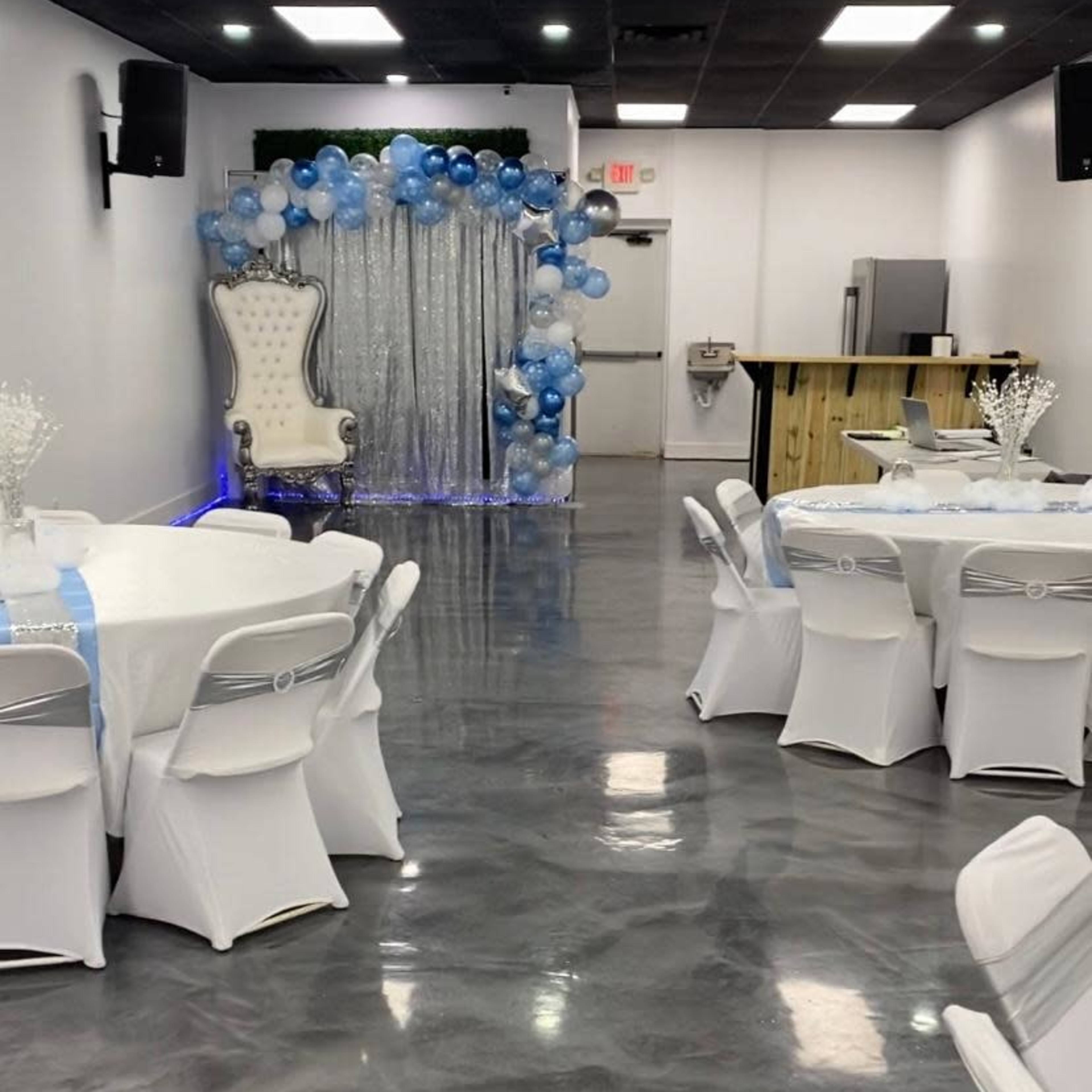 A decorated event hall with white tables, chairs, and a balloon arch in blue and silver behind a throne-like chair.