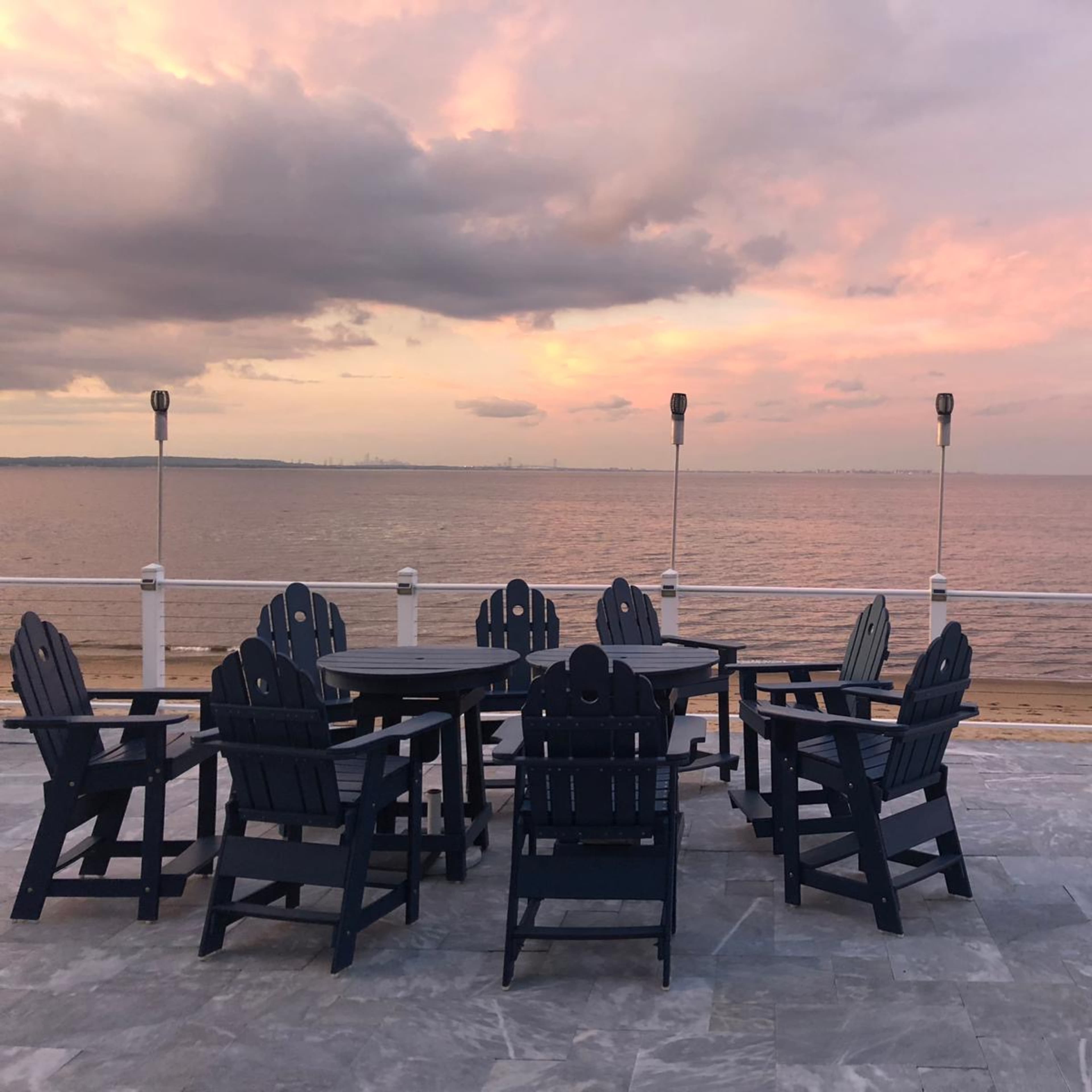 A row of dark wooden chairs and a table is arranged on a stone patio overlooking a calm sea at sunset.