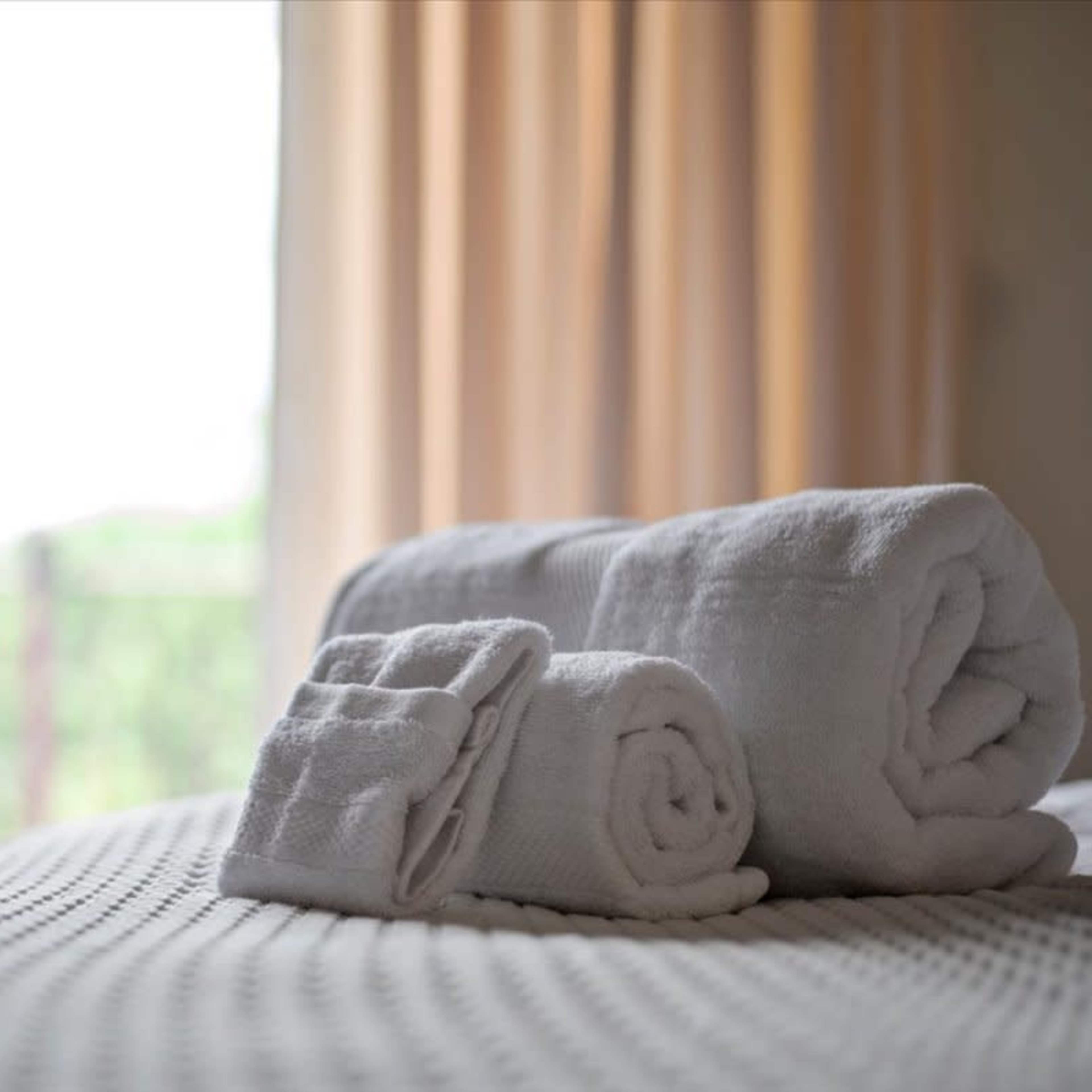 A stack of neatly rolled white towels is placed on a bed with a textured blanket, in a softly lit room.