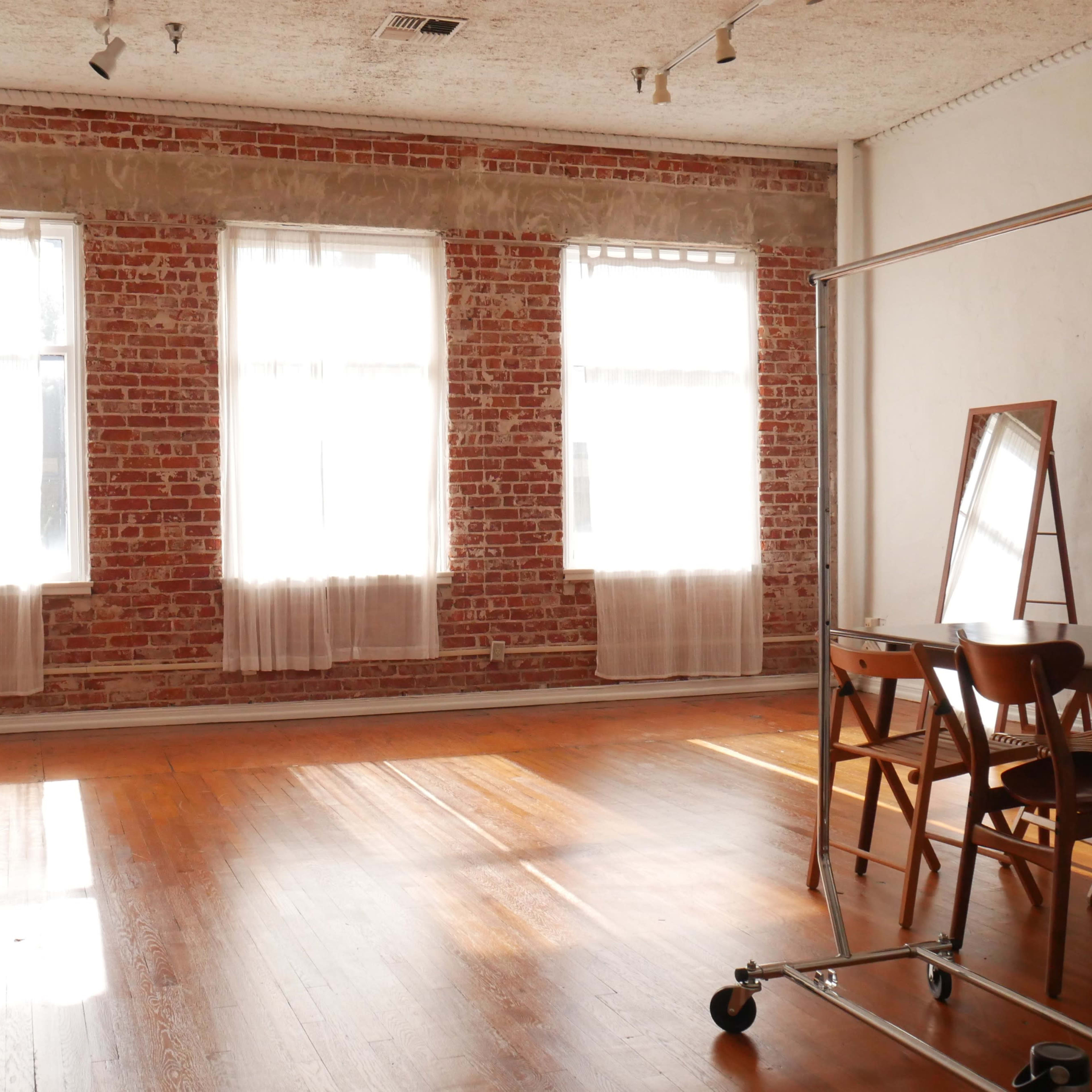 A spacious room with exposed brick walls, large windows, wooden flooring, and a table with chairs.