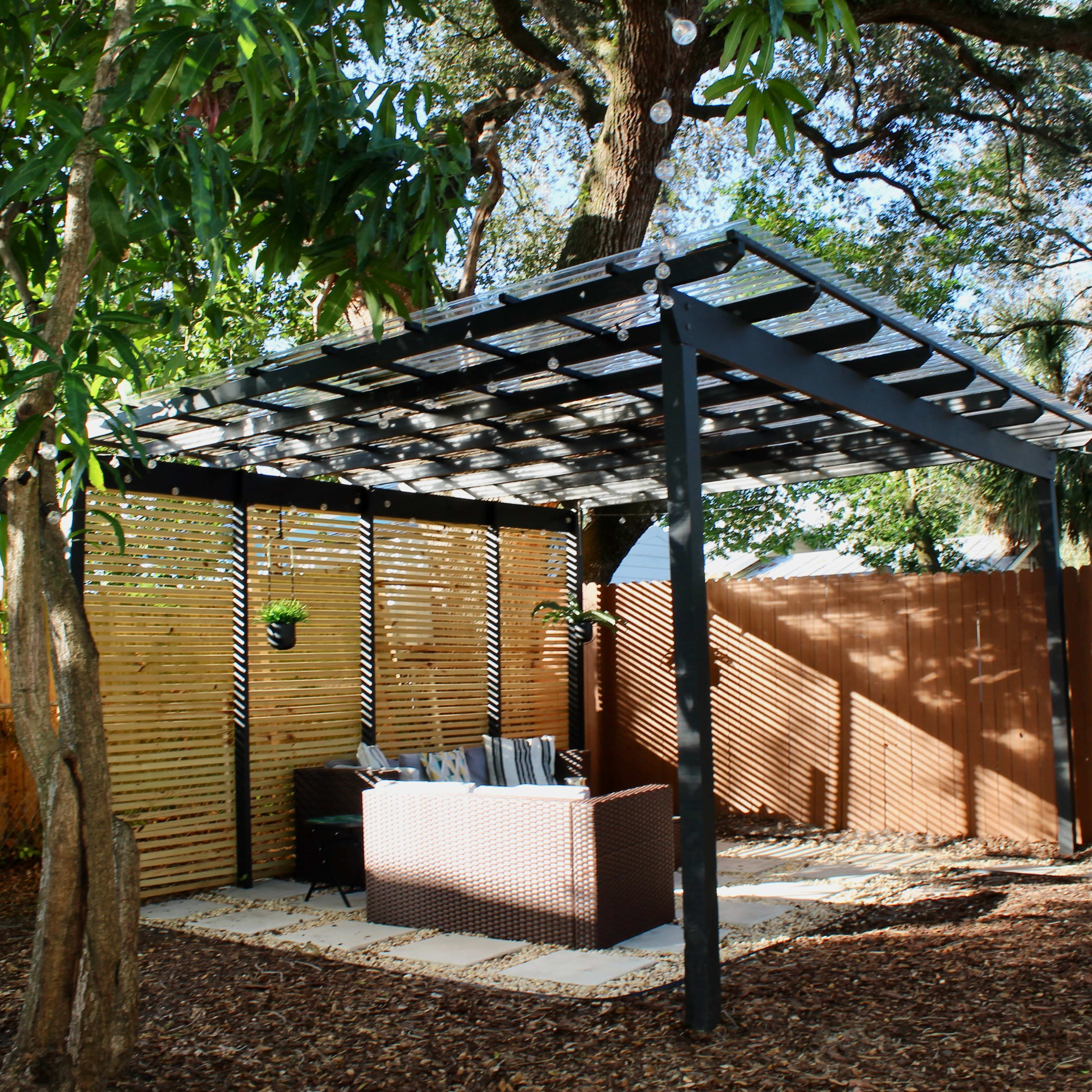 A shaded outdoor seating area with a pergola, surrounded by trees and a wooden fence.