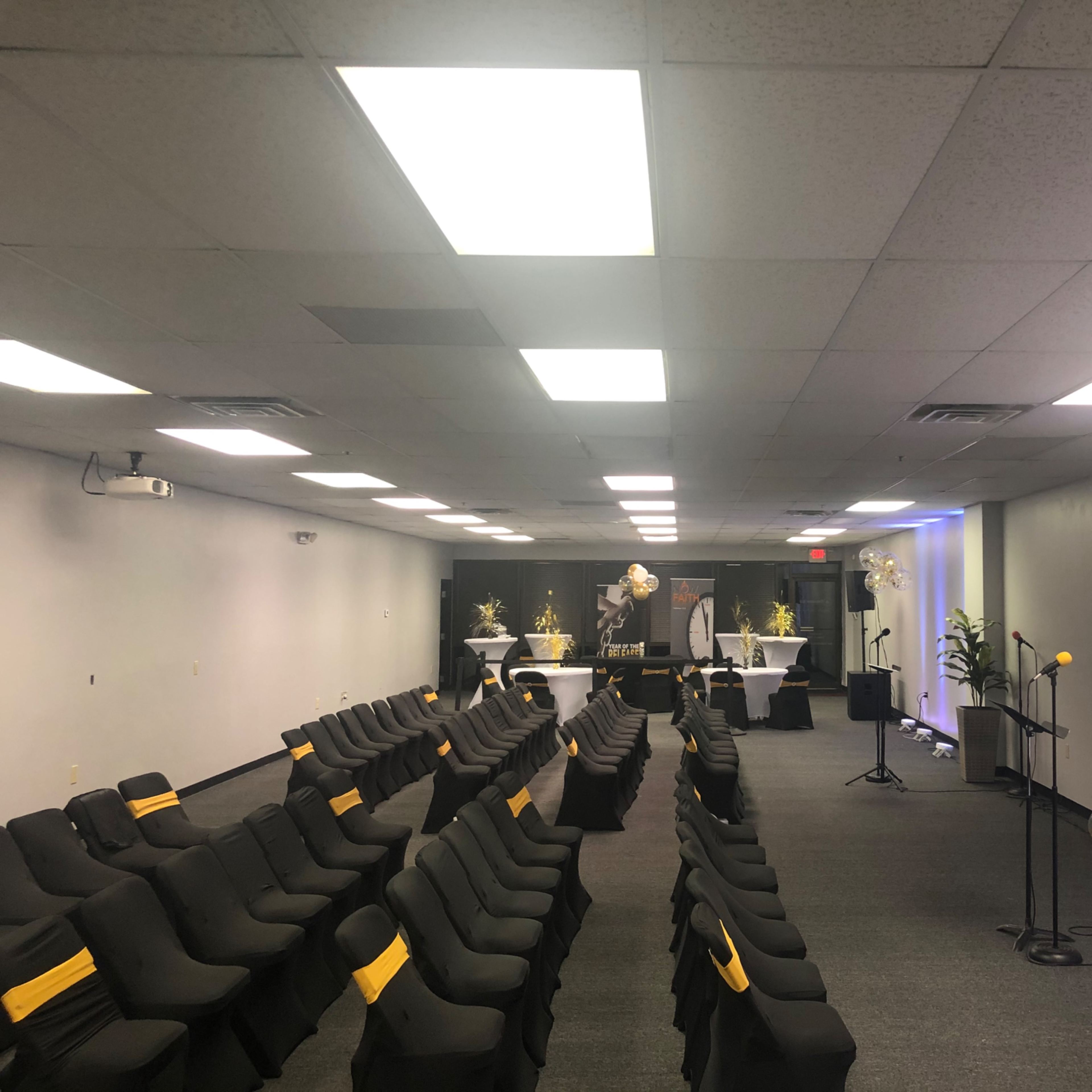 A modern event space set up with rows of black chairs featuring yellow accents, a stage with a microphone, and decorative elements including plants and tables.