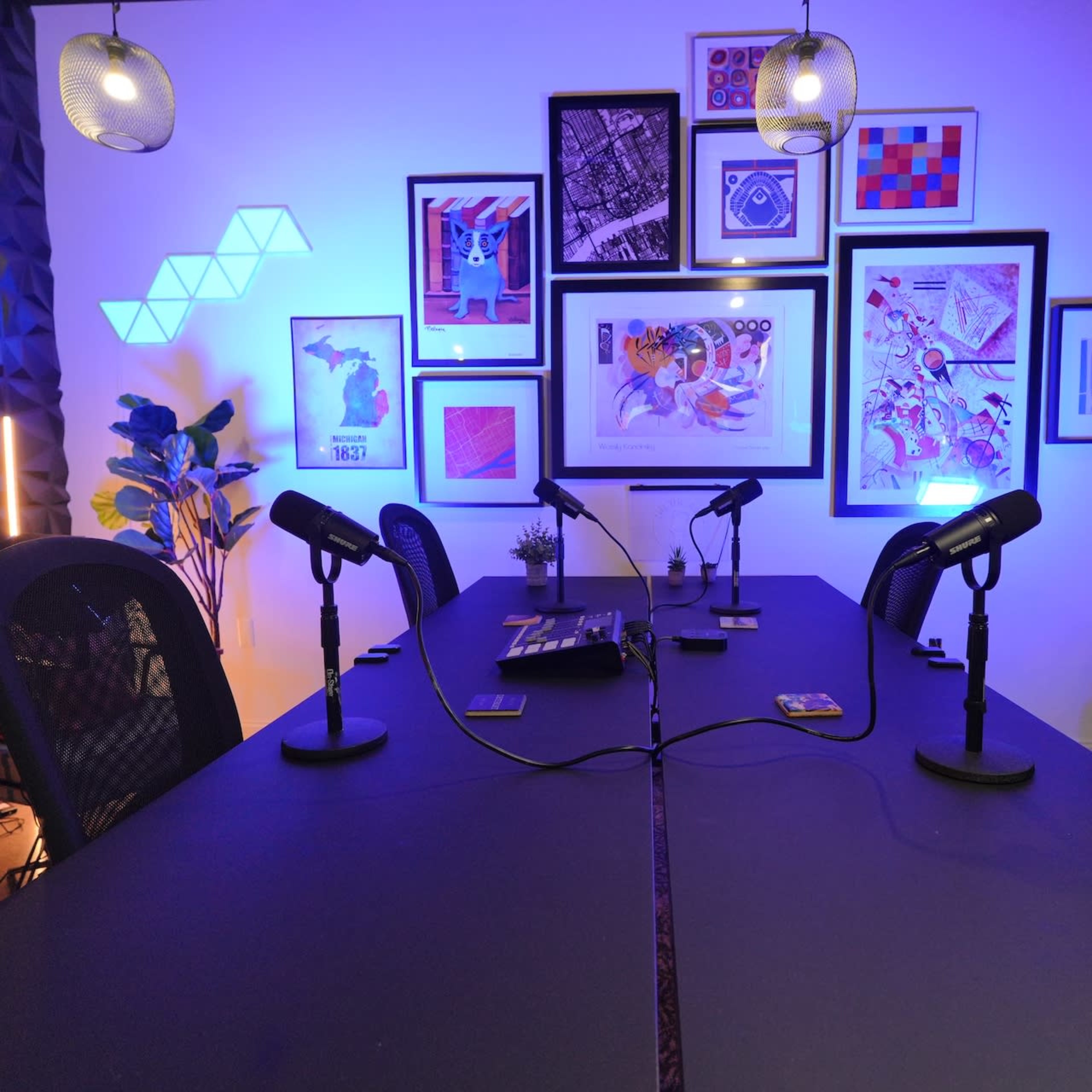A dimly lit podcast studio features a long table with microphones arranged on it and various framed artworks displayed on the wall.